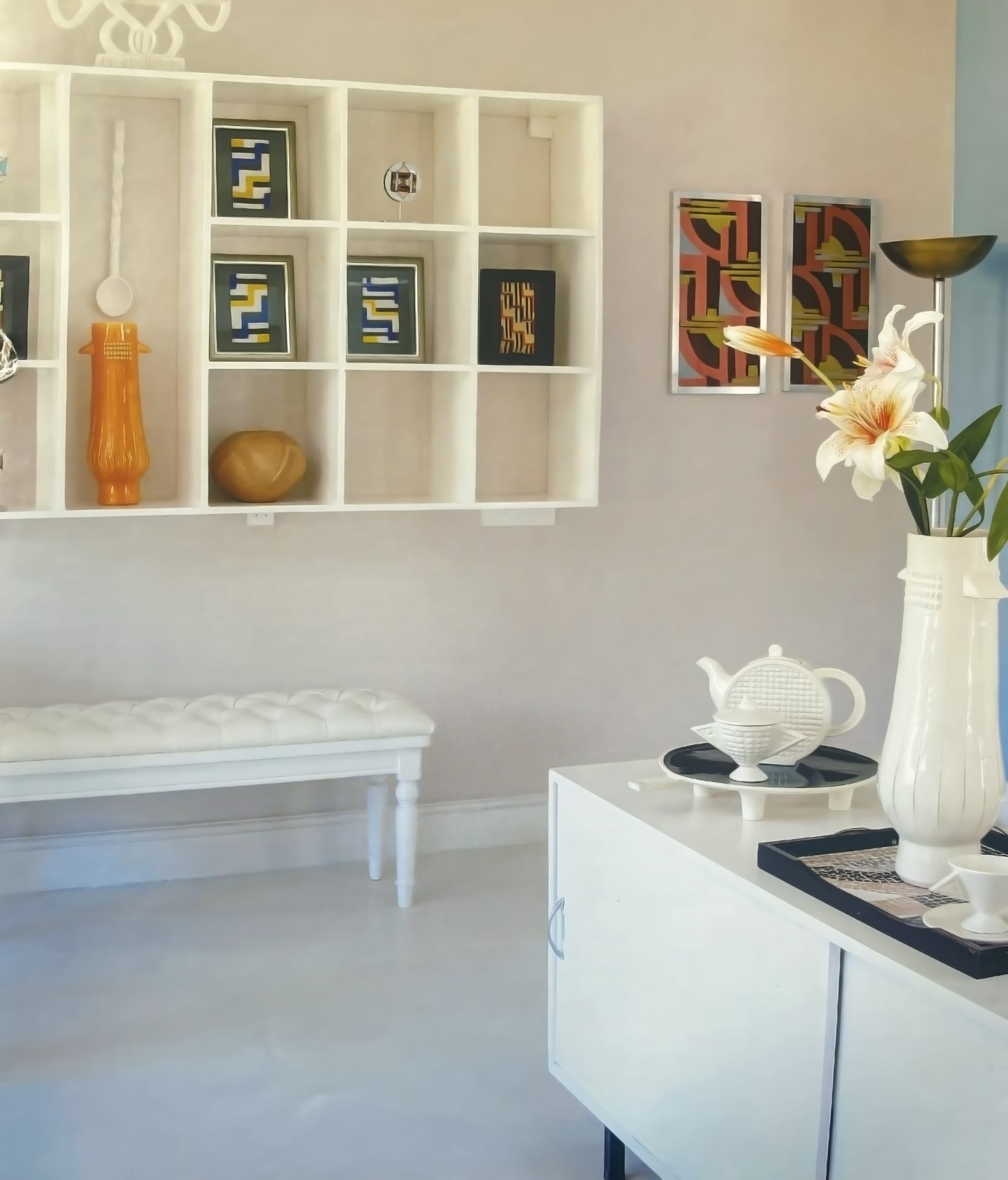 A modern living room with white walls, a wall-mounted white shelf displaying decorative vases and abstract art, a white padded bench, and a white sideboard topped with a tall vase of lilies, a teapot, and a tray.