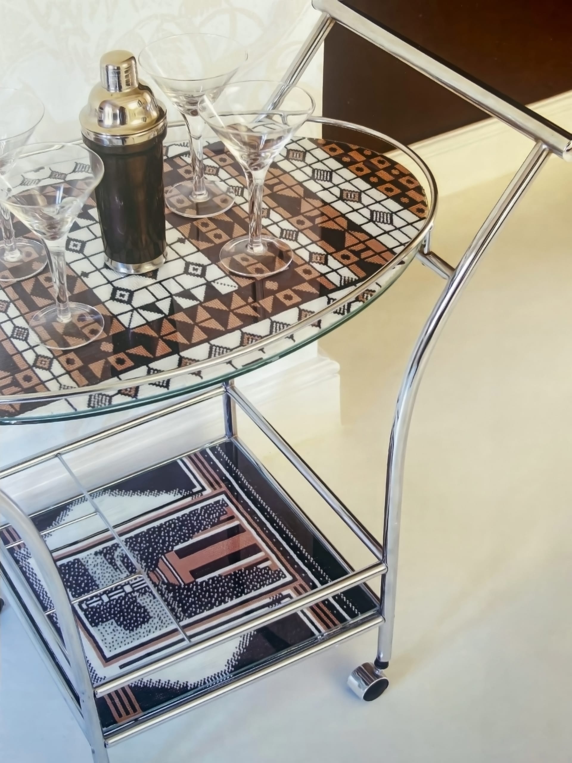 A chrome and glass drinks trolley stands on a cream floor. The trolley’s top shelf holds a silver cocktail shaker and four martini glasses on a geometric-patterned tray. Below, the lower shelf displays another pattern-covered tray with two chocolate bars.