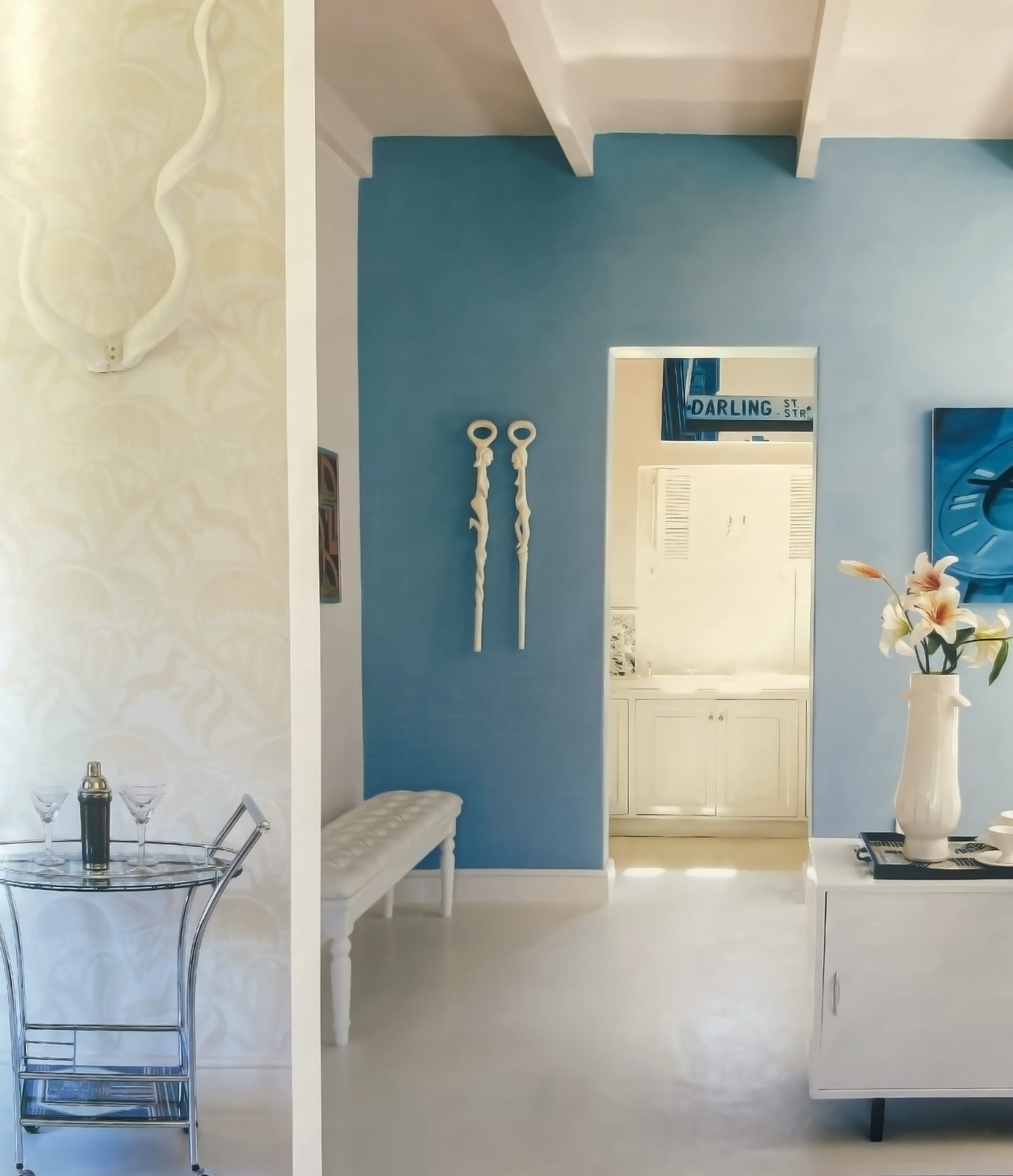 A modern living space with white floors and walls, a blue feature wall, a small bench, and decorative wall sculptures. A white cupboard with a vase of lilies is beside a drinks trolley with glasses. The room beyond has white cupboards and a “Darling” sign above the doorway.