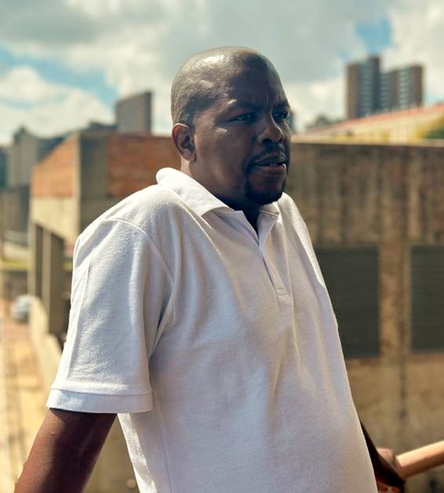 A man wearing a white polo shirt leans on a railing outdoors, looking to his left. Behind him are city buildings, a tall tower, and a cloudy blue sky, with cars and a street visible below. The lighting is bright and natural.
