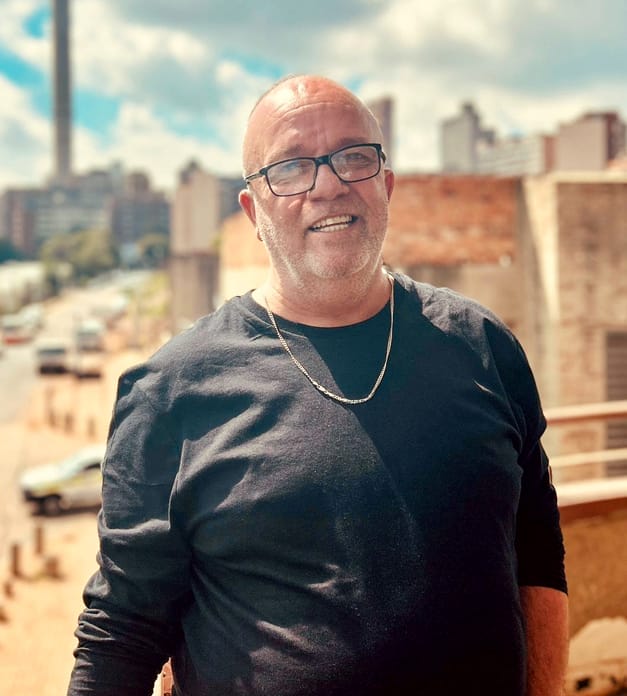 A smiling man with glasses, short hair, and a beard stands outdoors in sunlight. He wears a black long-sleeve shirt and a silver chain necklace. Behind him are brick buildings, a street with cars, and a city skyline under a partly cloudy sky.