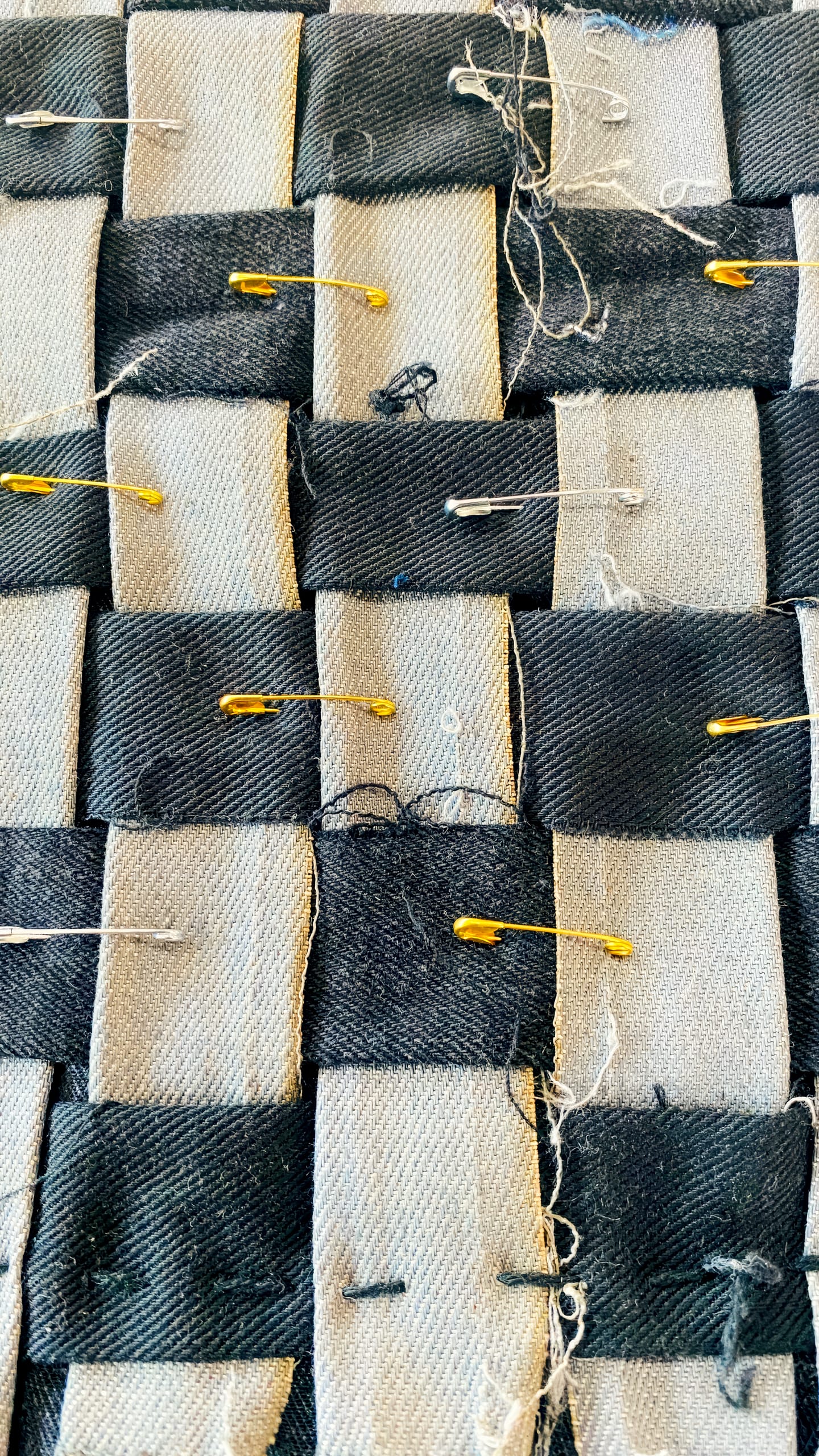 Close-up of woven black and light grey fabric strips with loose threads. Several gold and silver safety pins fasten the strips together. The woven pattern and rough textile texture are clearly visible in the image.