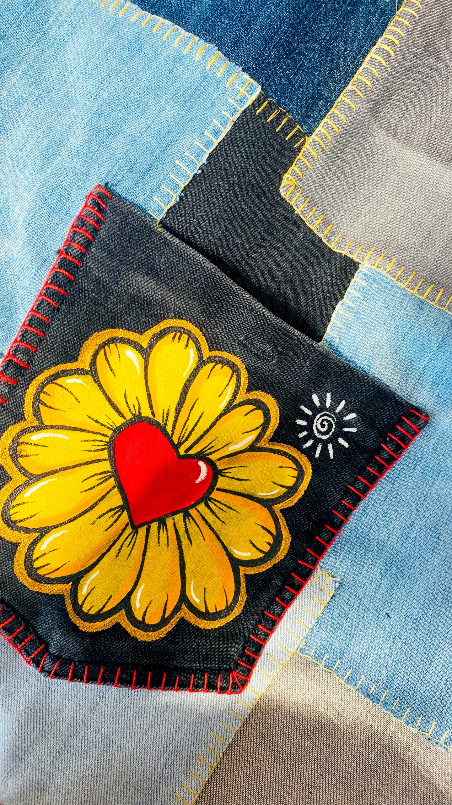 A denim patchwork with visible stitching features a pocket decorated with a hand-painted yellow flower with a red heart centre and a small white spiral, bordered by red thread. Various shades of blue and grey fabric pieces surround the pocket.
