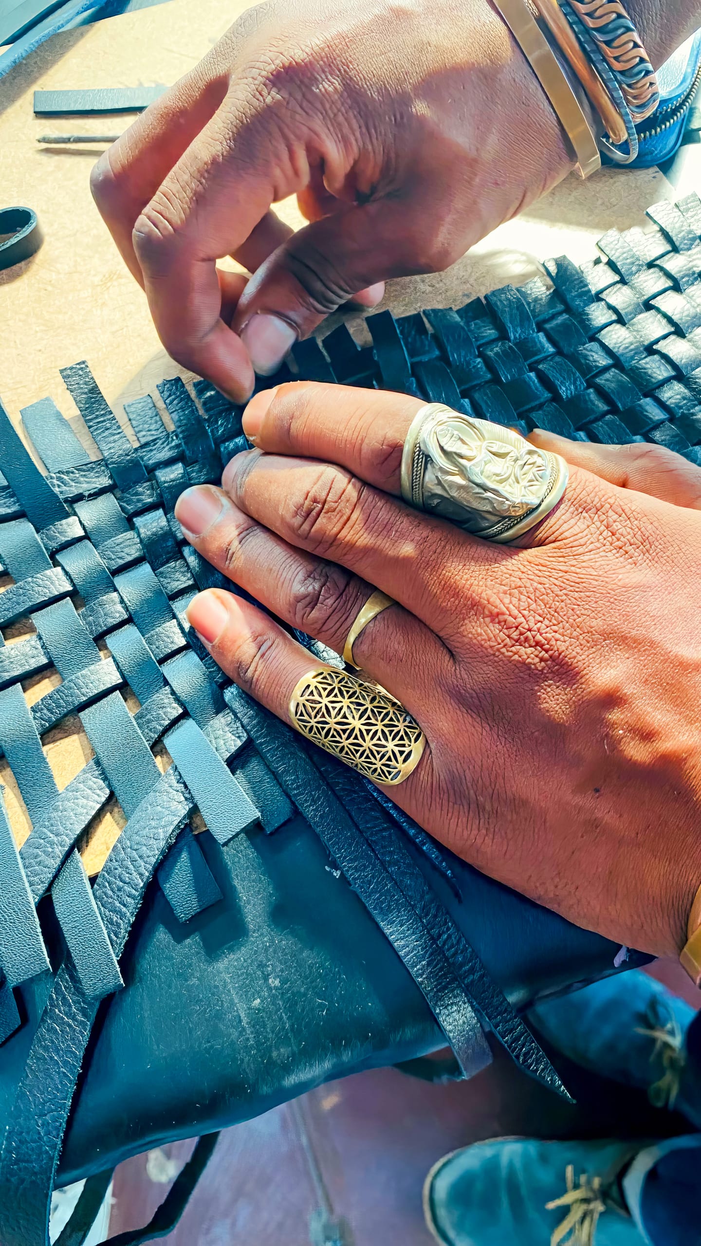 Close-up of hands weaving strips of black leather. The person wears three ornate rings and a bangle. Sunlight highlights the texture of the leather and the jewellery’s intricate patterns, emphasising the craftsmanship in the weaving process.