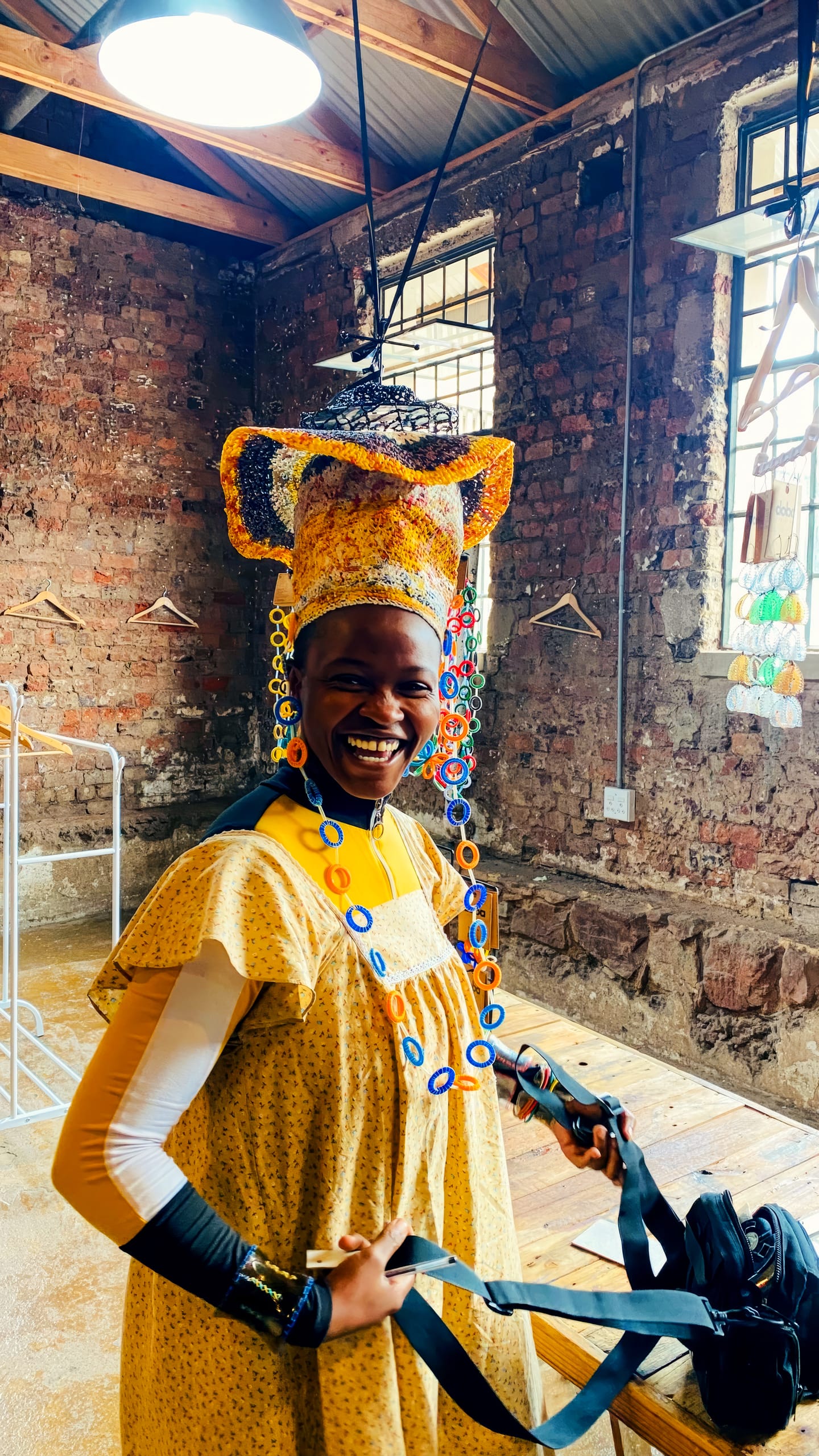 A smiling person in a yellow patterned dress and elaborate yellow and blue beaded headdress stands indoors near rustic brick walls and windows, holding a black strap. Sunlight streams in, illuminating their joyful expression and vibrant outfit.