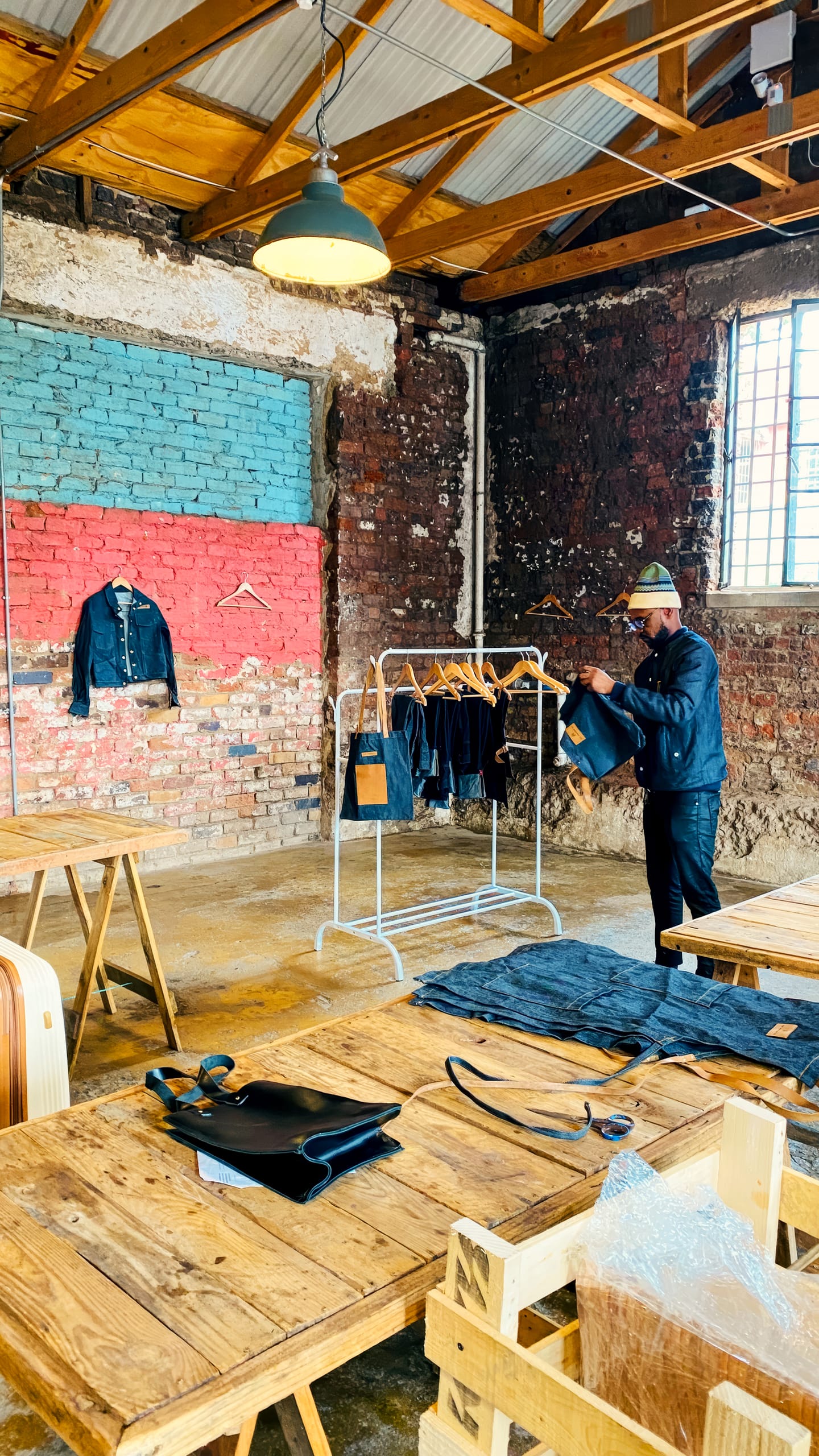 A person arranges denim clothing on a rail in a rustic room with exposed brick walls, wooden tables, and a large window. Jackets and bags are displayed, and the space has an industrial, creative atmosphere with visible ceiling beams.