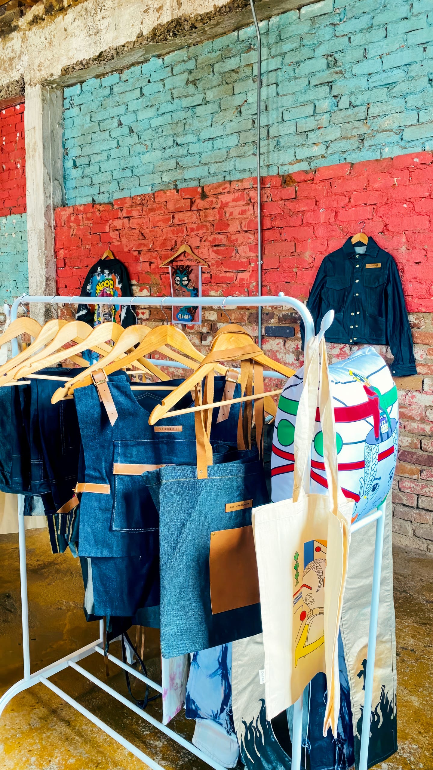 A clothes rail displays denim jackets, aprons, and tote bags with leather accents. One colourful canvas tote with graphic art hangs at the front. Behind, a brick wall painted blue and red and a concrete floor create an industrial-style backdrop.