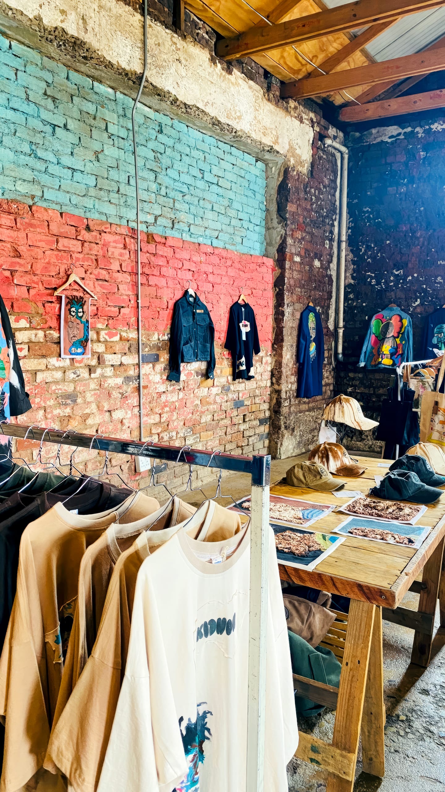 A rustic shop with exposed brick walls displays colourful shirts and jackets on hangers and rails. Wall art and framed pieces hang on a turquoise and red-painted section. Wooden tables hold prints, accessories, and folded clothes under slanting sunlight.