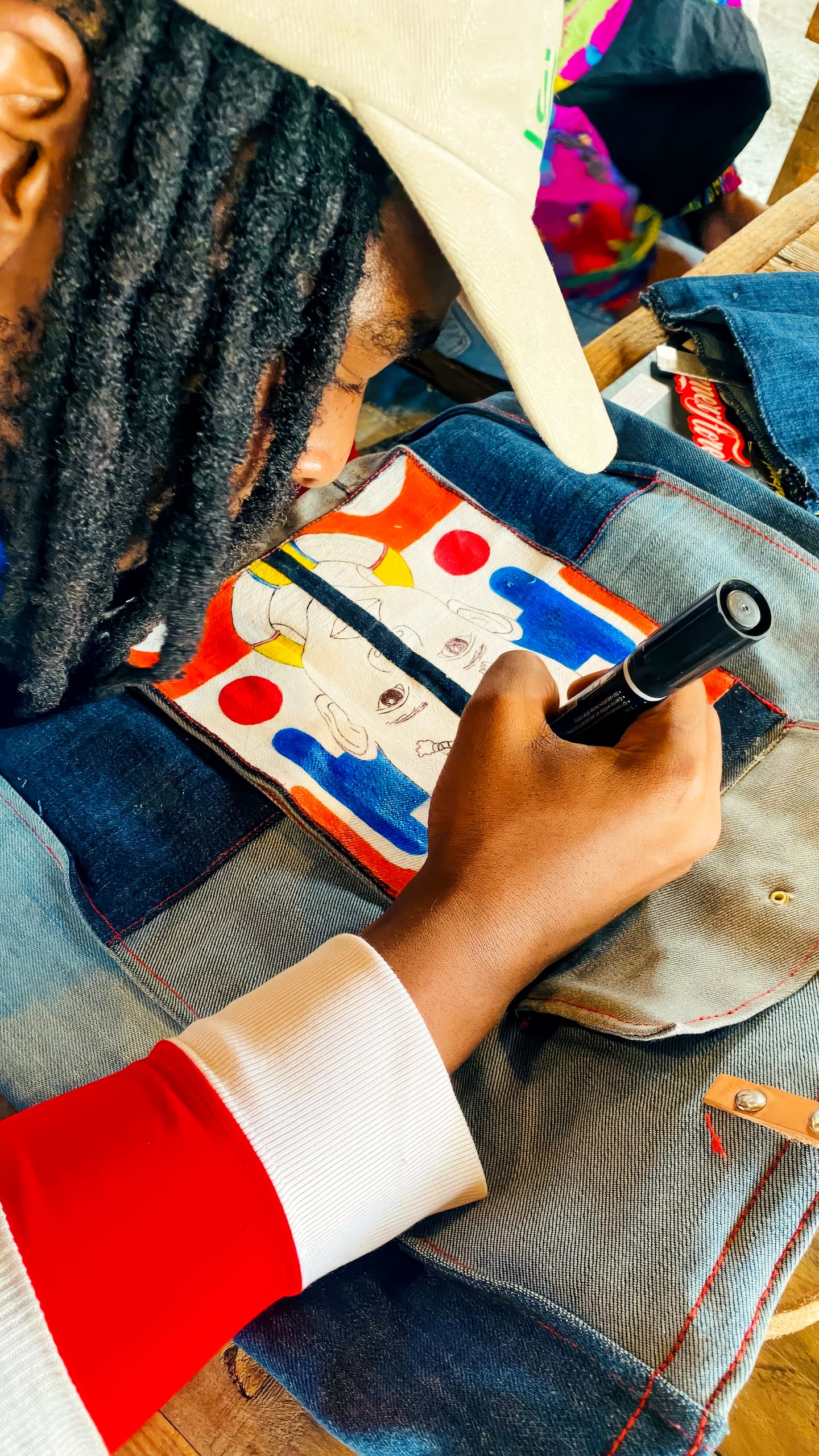 A person with long hair and a tan cap draws on denim fabric using a black marker. The fabric features a colourful artwork with red, blue, and yellow shapes, and two faces. The person wears a red and white striped sleeve.