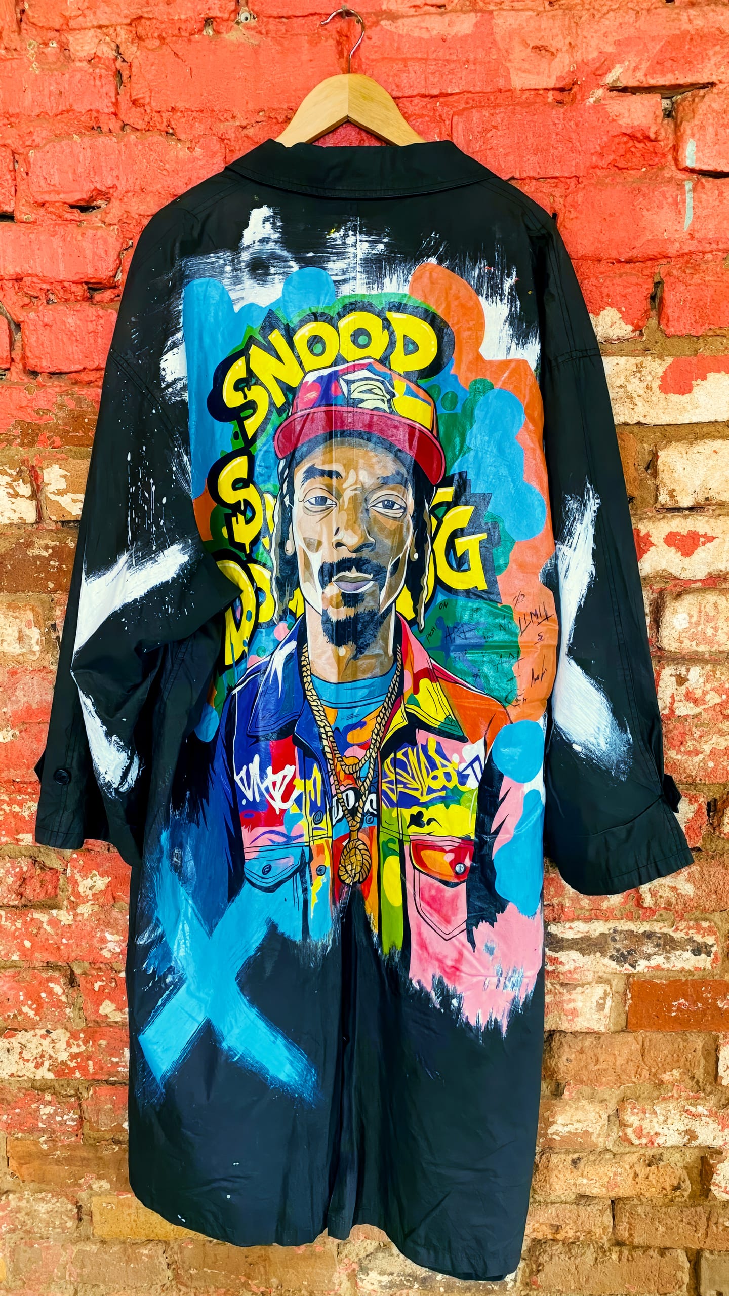 A long black coat hangs against a weathered red brick wall. The back features a colourful, hand-painted portrait of rapper Snoop Dogg in bright clothing and a red cap, with his name “SNOOP” in bold yellow letters above and graffiti-style accents around him.