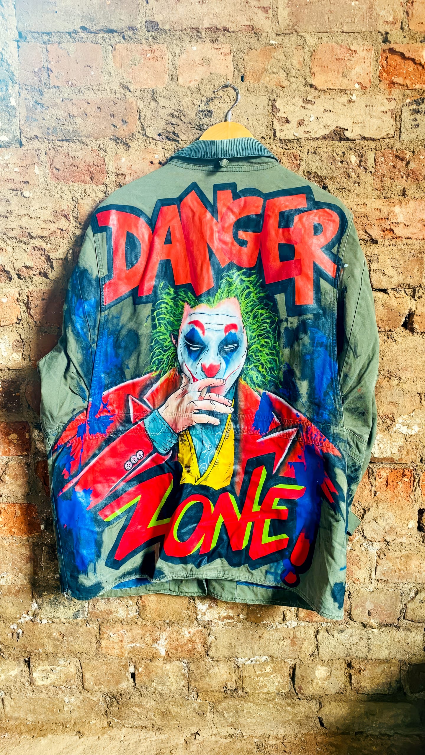 A painted denim jacket hangs on a brick wall. The back features a vivid illustration of a Joker-like character with green hair, make-up, and a red suit, smoking with two fingers. Bold red letters read “DANGER ZONE!” above and below the character.