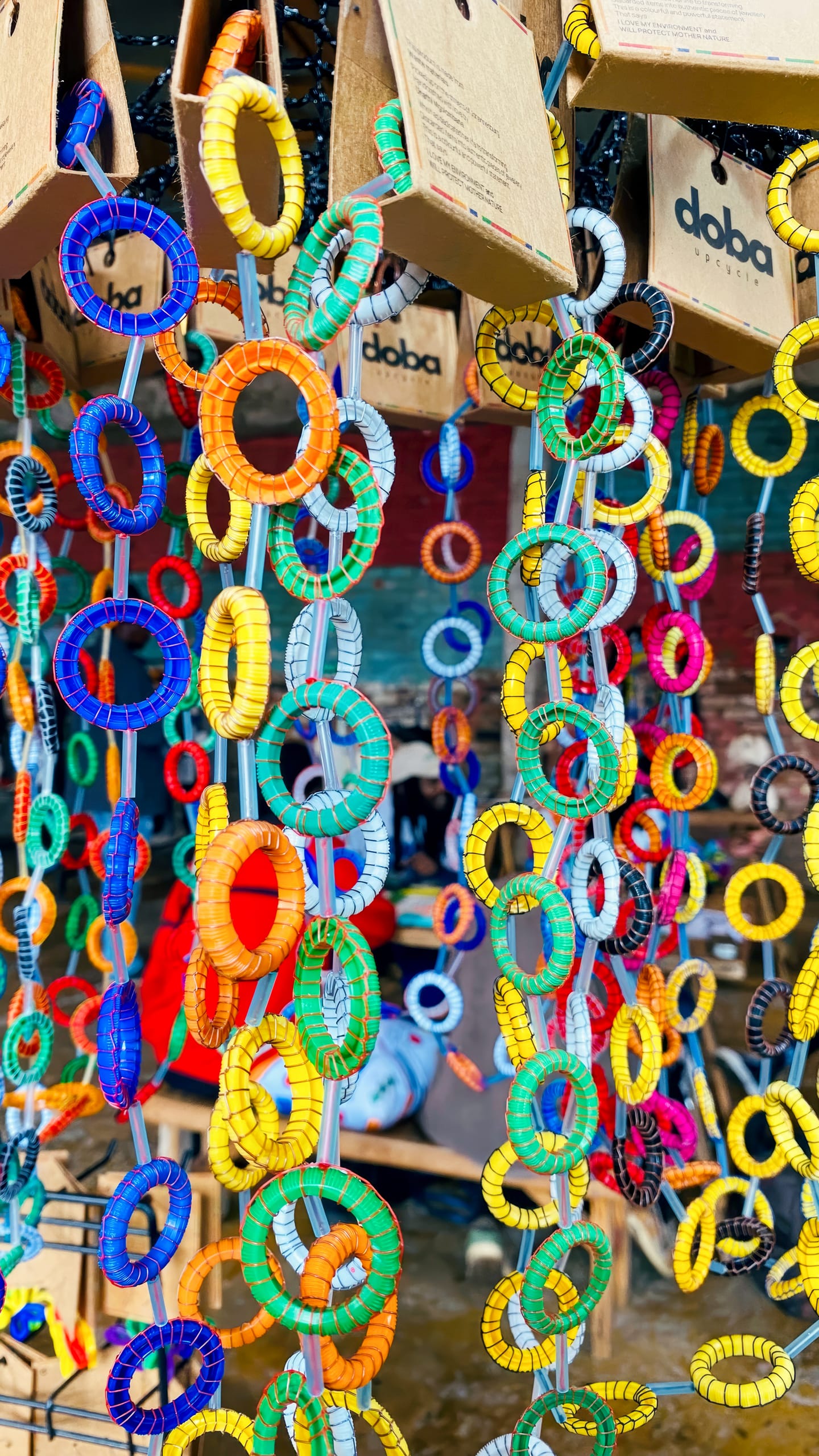 Colourful, circular plastic rings are strung together to form long, hanging chains displayed at a market stall. The rings are in bright shades of blue, yellow, orange, green, red, and white. Cardboard tags hang above with the brand name 