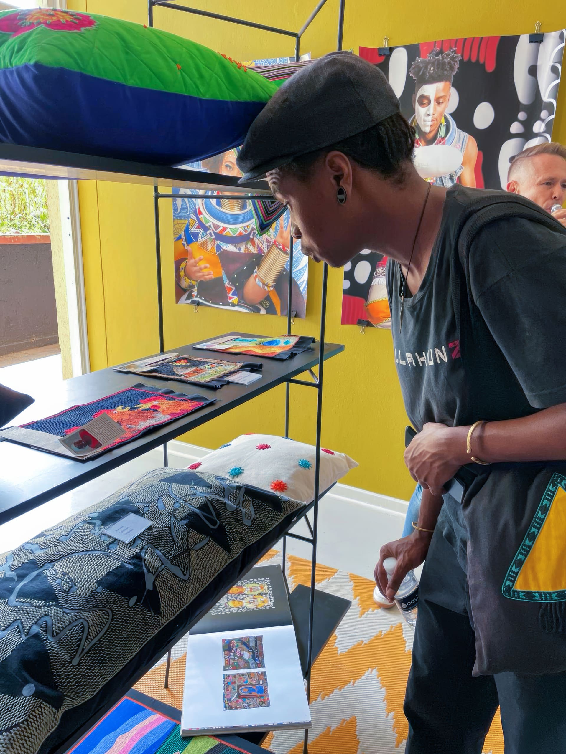 A person wearing a cap and black shirt leans forward to closely examine colourful, patterned textile items displayed on shelves. Artworks and cushions with vibrant designs are visible. Another person stands partly visible in the background against a yellow wall.