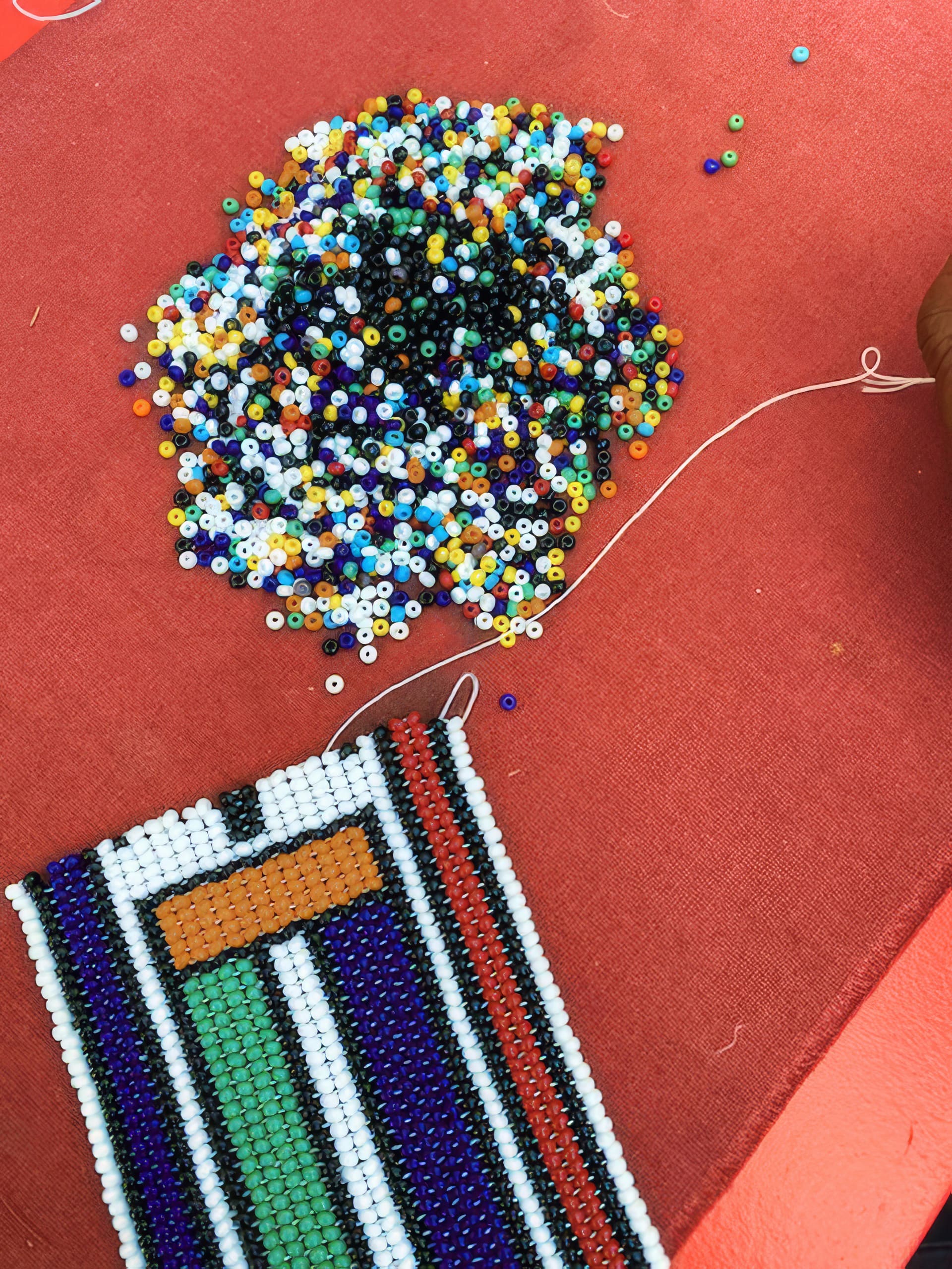 A beaded bracelet is being made on a red surface. Multicoloured loose beads are scattered above, while the partially finished bracelet features geometric patterns in black, white, green, red, blue, and orange beads. A white thread is visible, connecting the beads.