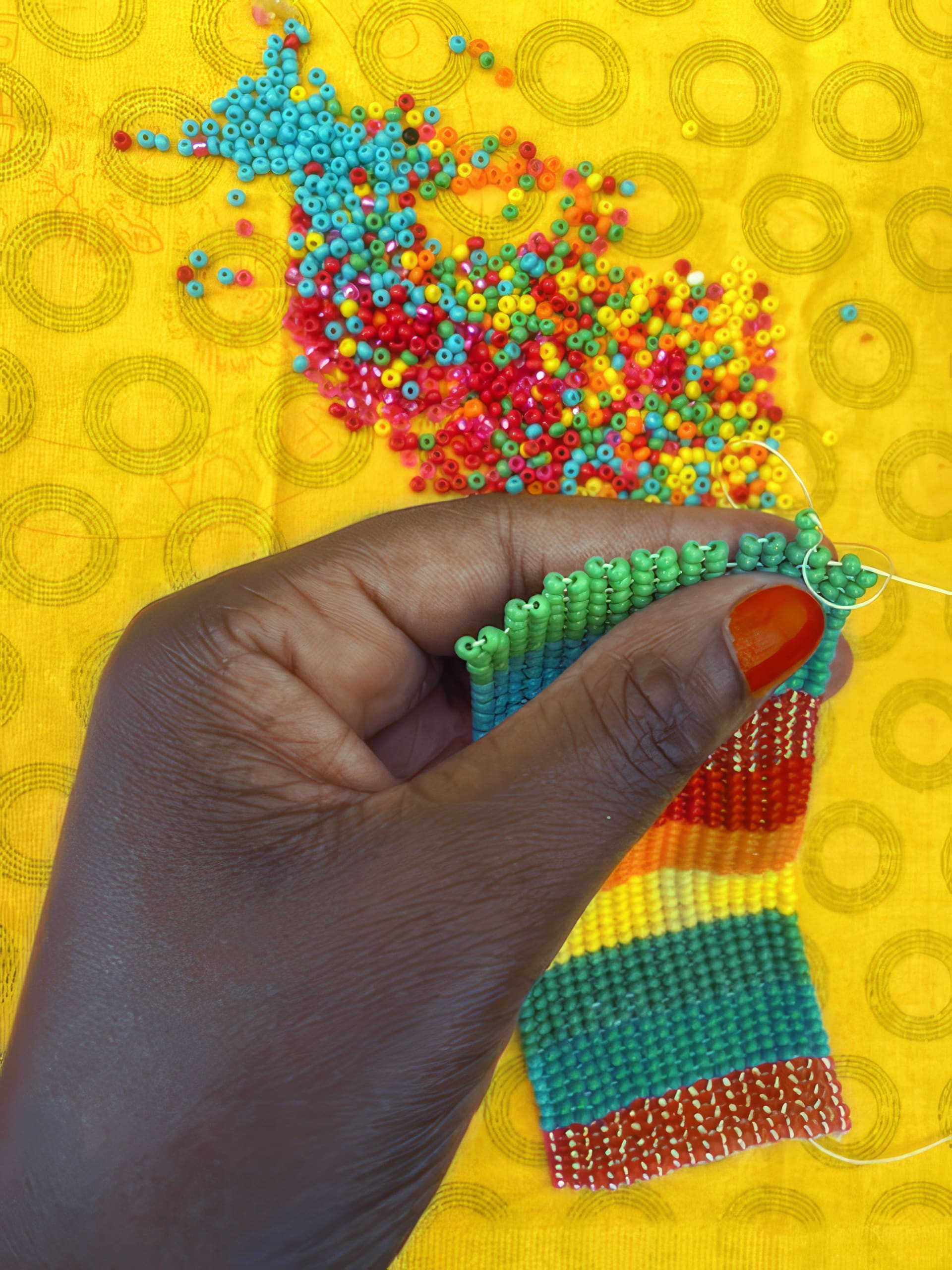 A close-up of a hand with bright orange nail varnish weaving a colourful beaded bracelet. The beads are green, yellow, red, and blue, and are scattered on a yellow patterned fabric background. The hand is threading green beads onto the bracelet.