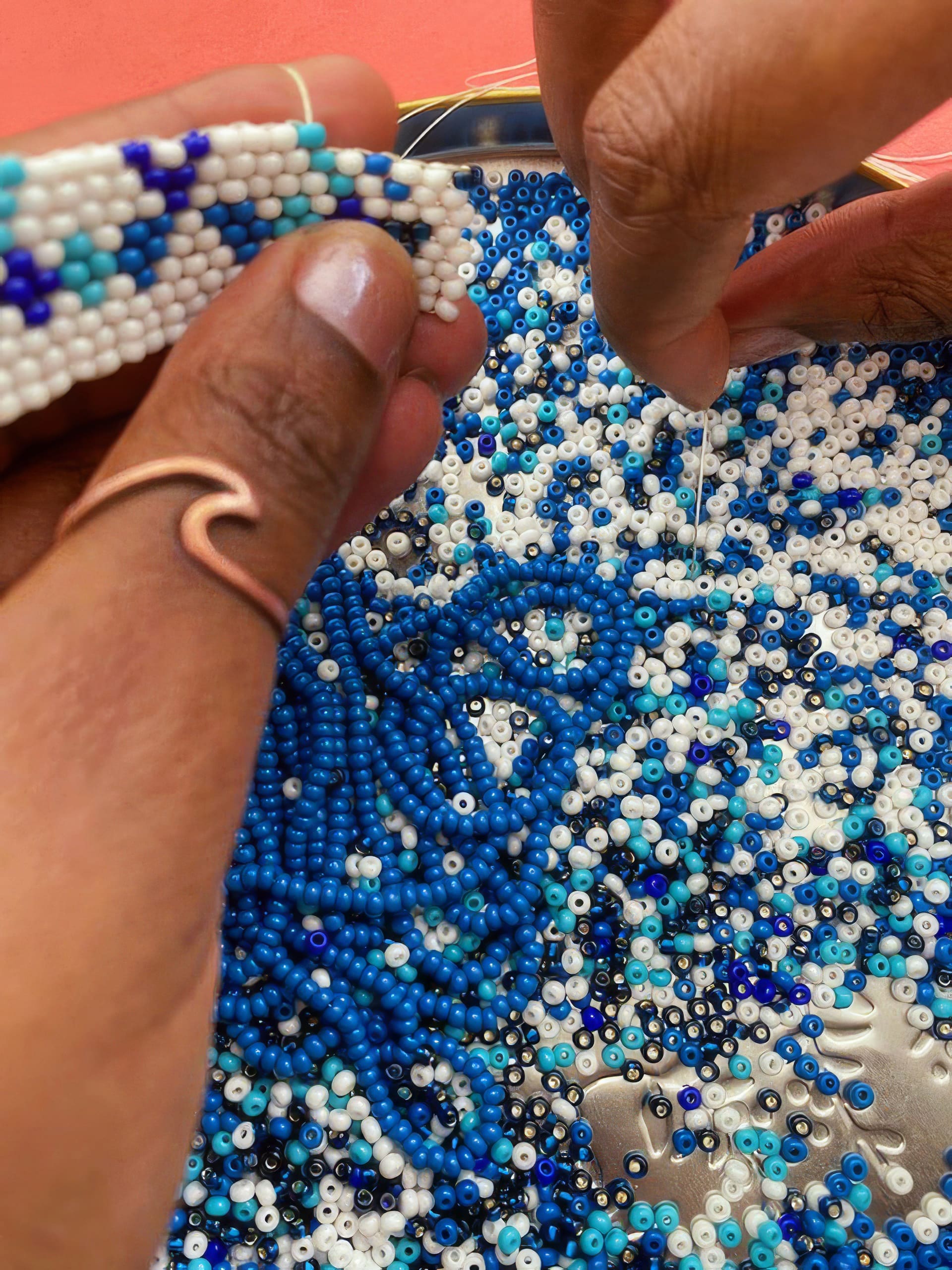 Close-up of hands threading blue, white, and turquoise beads onto a string above a tray filled with multicoloured beads. One hand wears a copper wave-shaped ring, and a woven bead strip is being created against a red background.