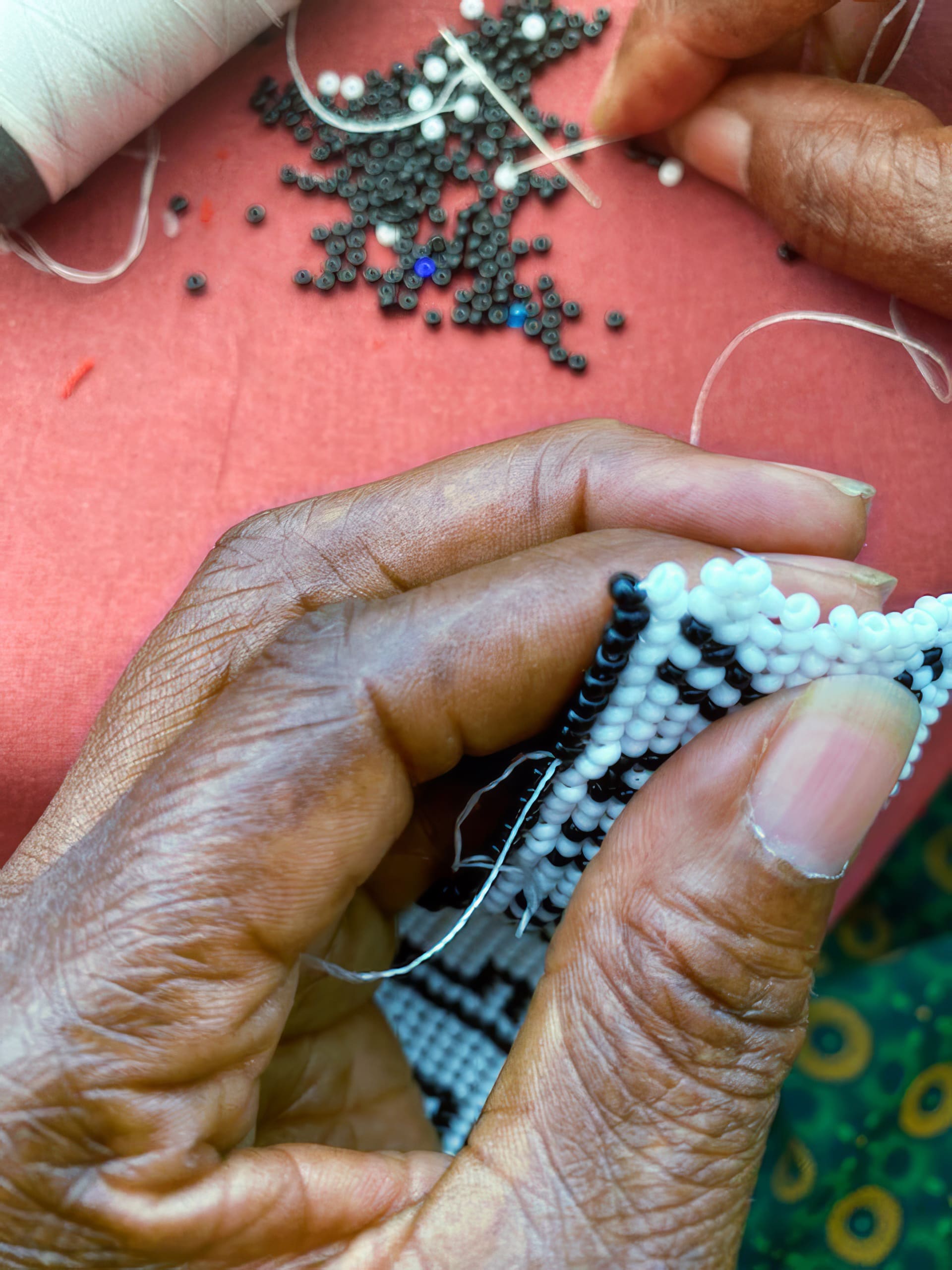 A close-up of hands sewing black and white beads onto fabric. A reel of white thread, a needle, and scattered loose beads are visible on a pink surface in the background. The person's skin is dark, and their green patterned clothing is partly visible.