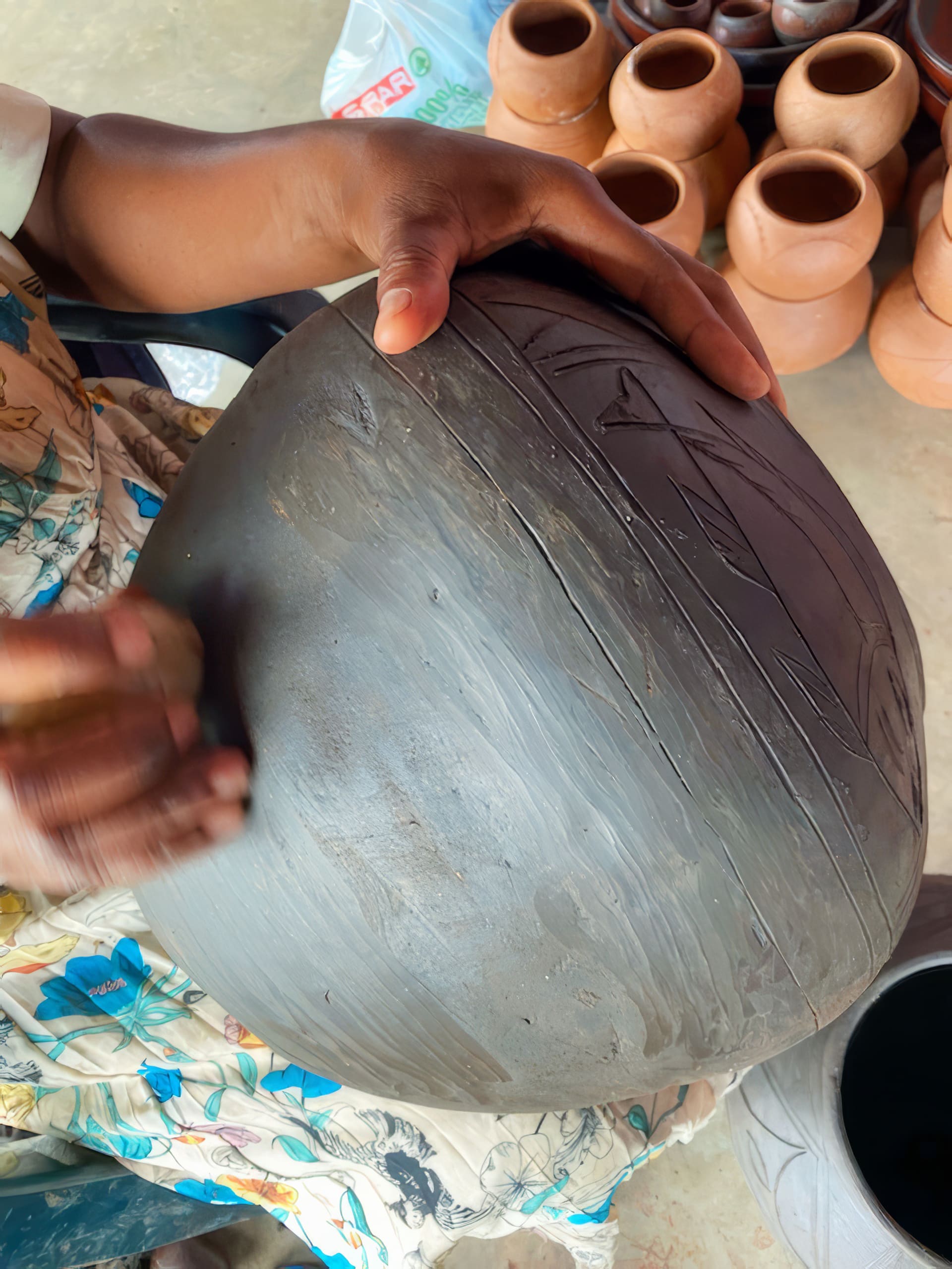 A person’s hands are carving decorative lines into a large, round, dark clay pot. They are using a small tool on the surface. Several finished terracotta pots are visible in the background on a concrete floor. The person wears a floral-patterned outfit.