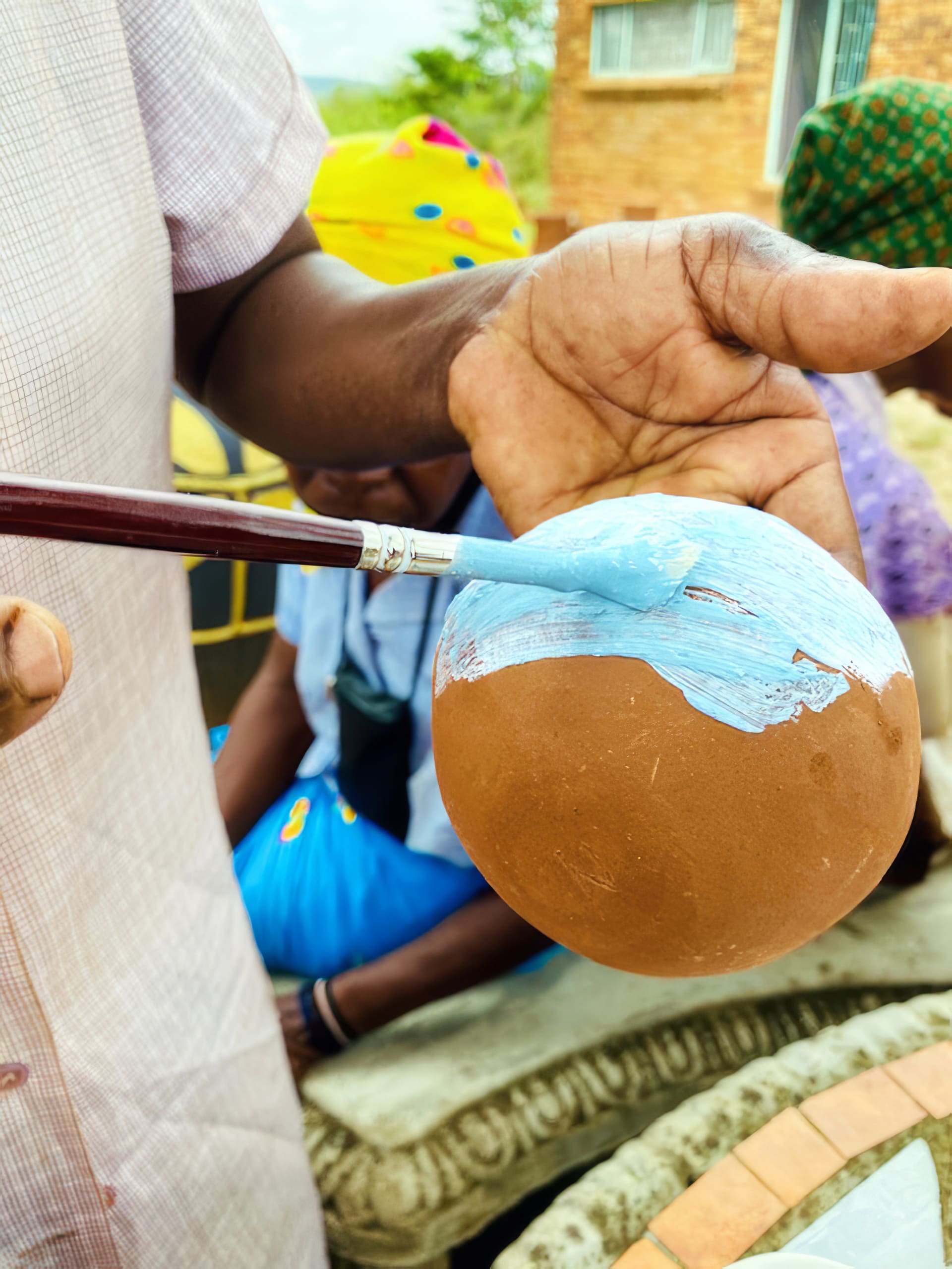 A close-up of a person’s hand holding a round, brown object while using a paintbrush to apply light blue paint. In the background, people in colourful clothing and headwraps sit outdoors near a brick building. The focus is on the painting activity.