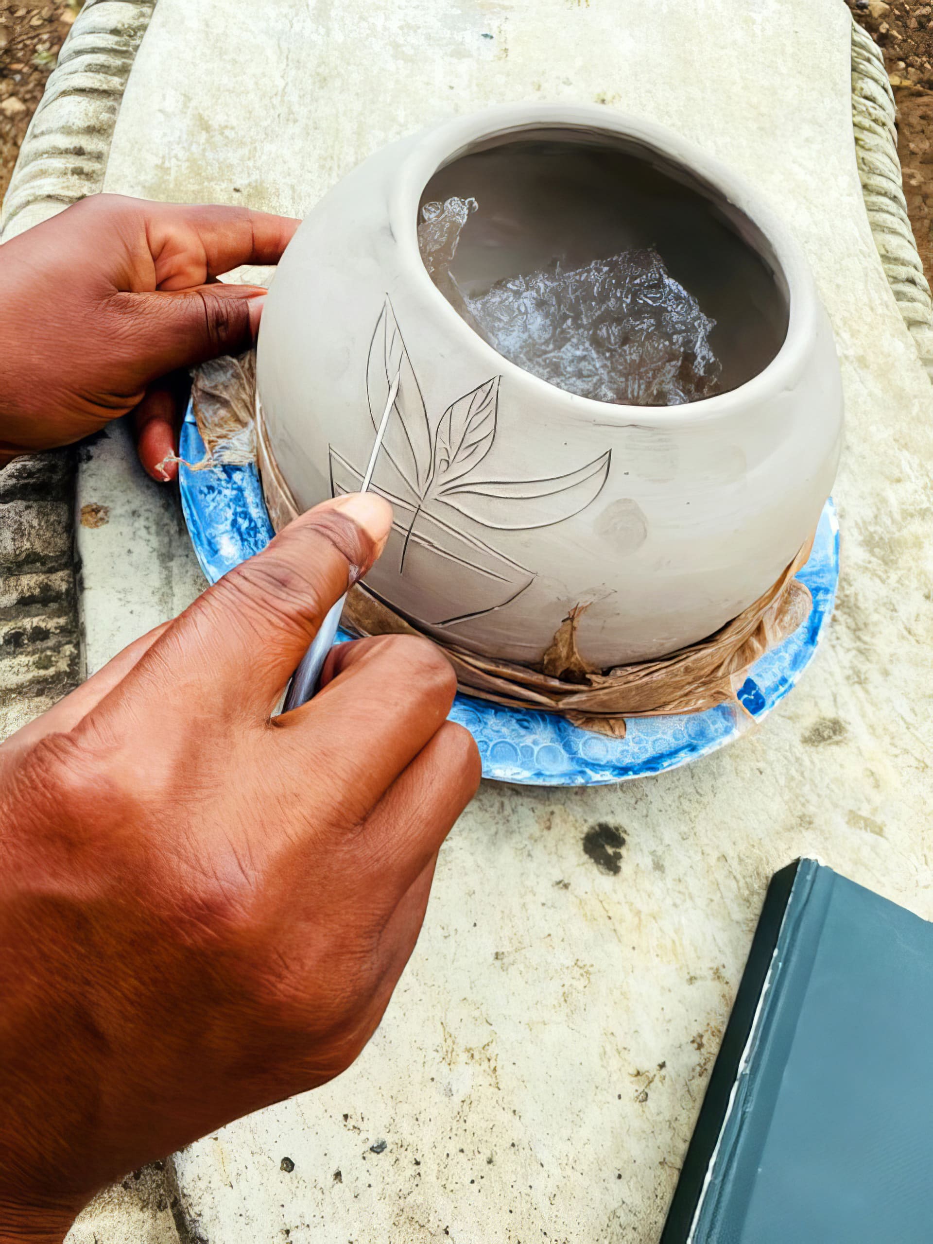 A person uses a metal tool to carve a leaf design onto the side of a round, unfinished ceramic pot placed on a blue plate. The pot sits on a white surface, and a green book is visible in the lower right corner.