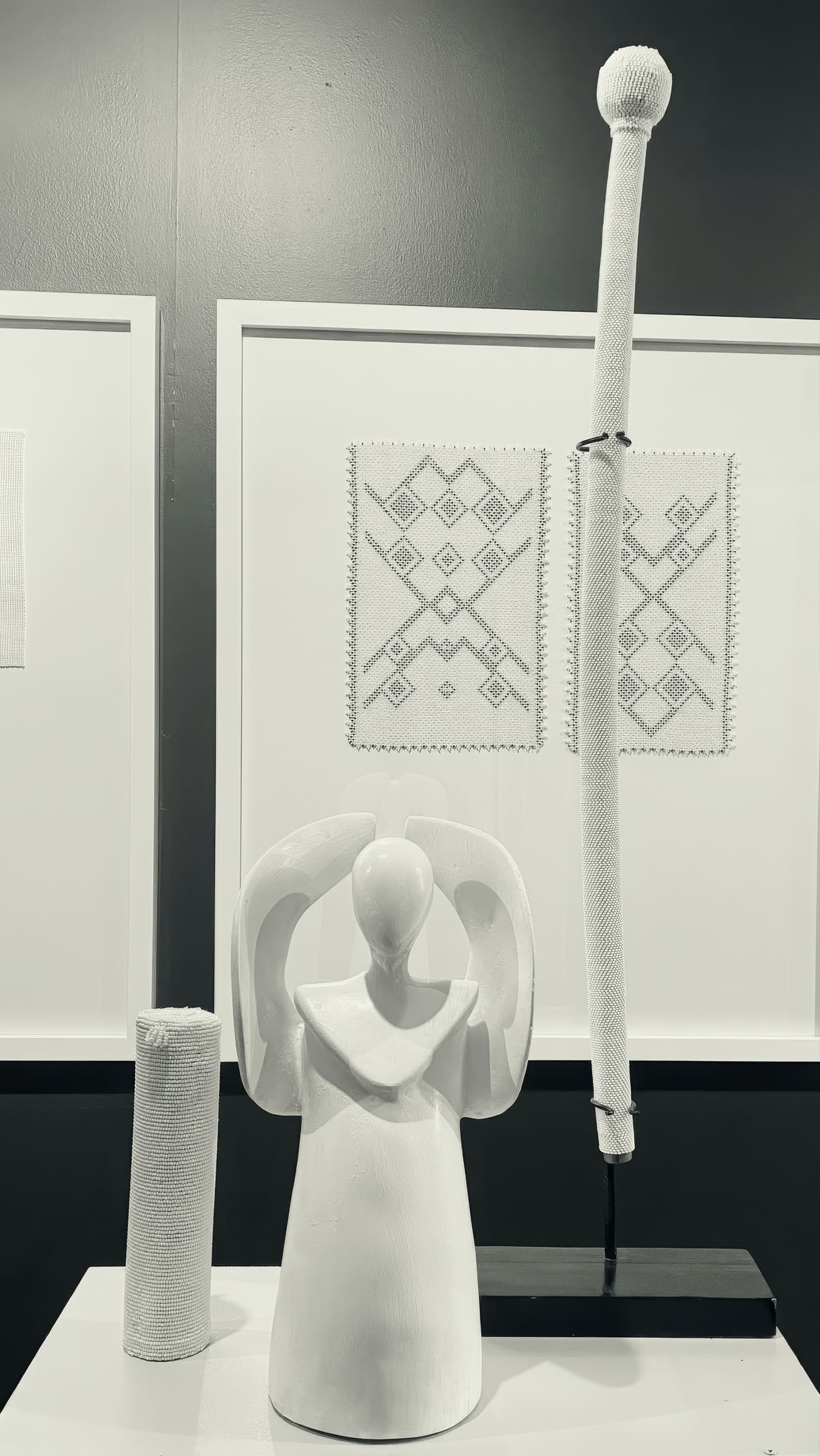 A black-and-white photo shows a smooth, abstract angel sculpture with arms raised. Behind it are two geometric embroidered fabric pieces framed on the wall, and to the right stands a tall, wrapped staff with a textured sphere on top.