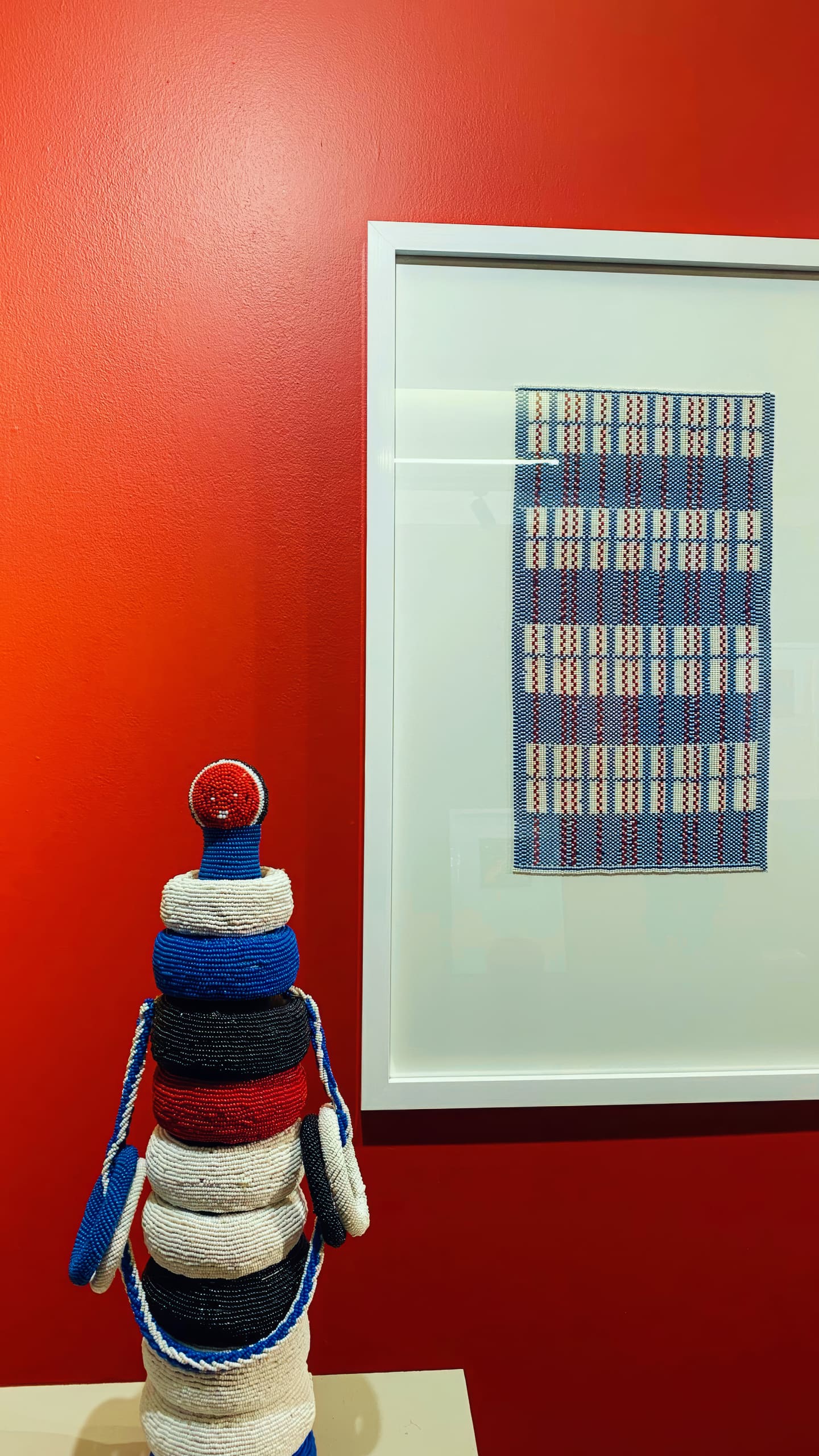 A colourful sculpture made of stacked, woven rings in red, white, blue, and black sits on a white surface against a vivid red wall. To the right, a framed artwork displays a blue, white, and red rectangular woven pattern.