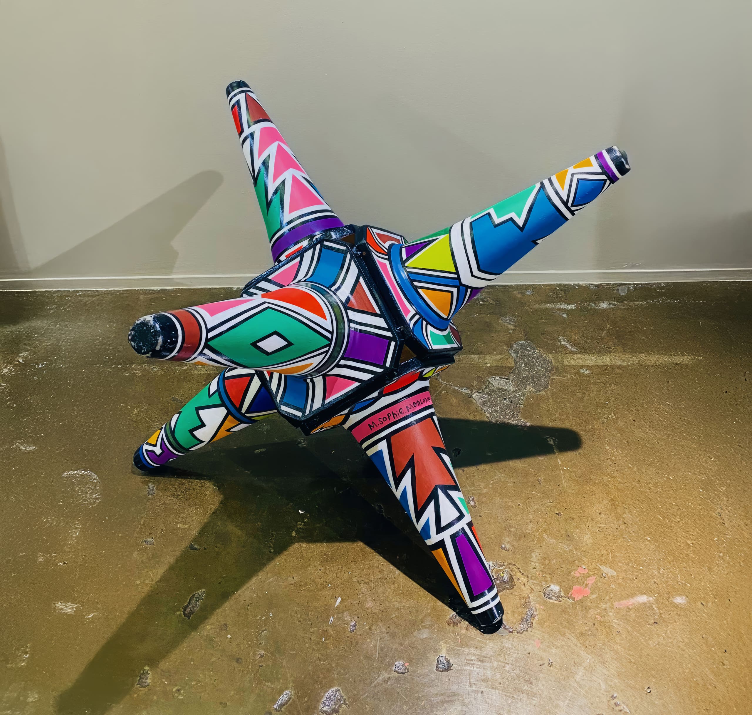 A colourful, four-pointed geometric sculpture with bold, angular patterns in red, blue, green, purple, and black sits on a polished brown concrete floor against a plain beige wall. Each point is uniquely decorated with vibrant zigzags and shapes.