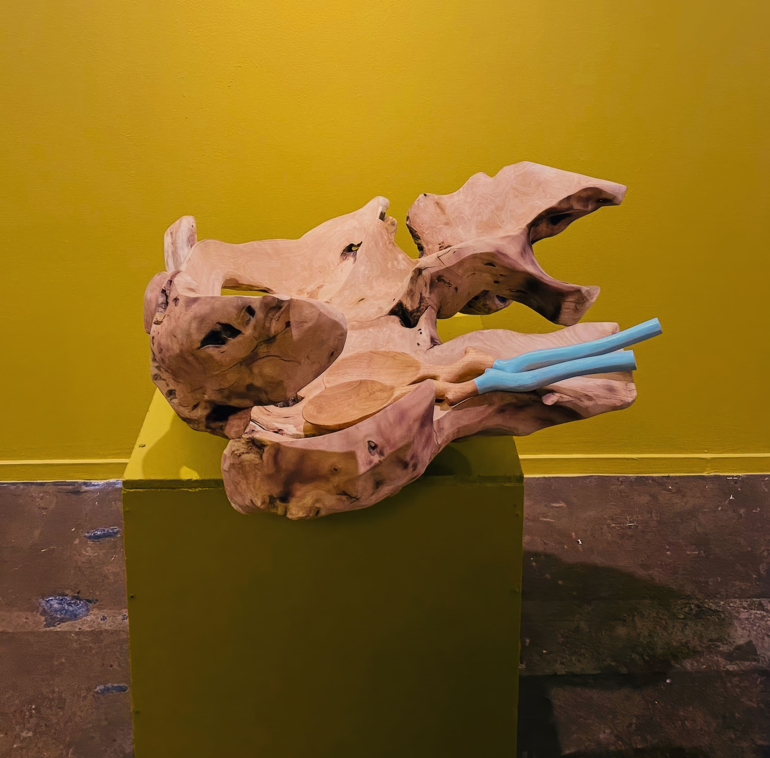 A pale, organic-shaped wooden sculpture with twisting forms rests on a green plinth. Two blue sculpted chopsticks lie on part of the wood, against a yellow wall and a mottled brown stone floor.