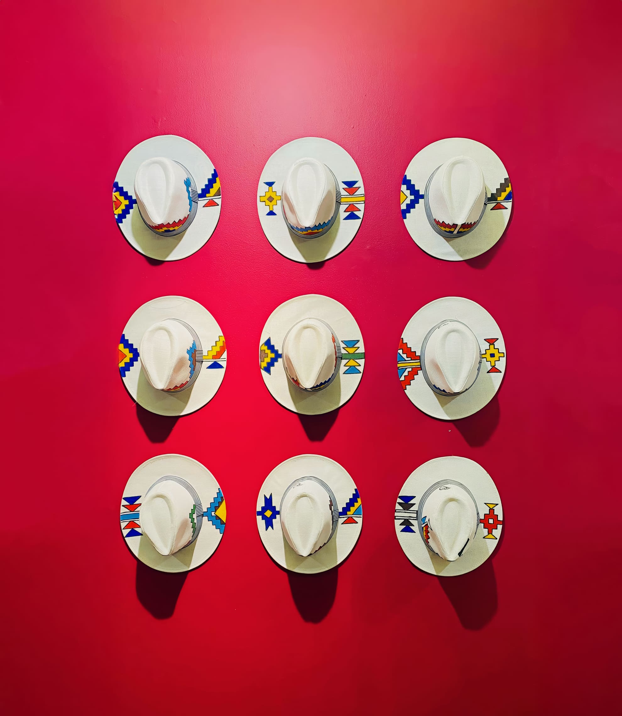 Nine white cowboy hats with colourful beaded hatbands are mounted in a three-by-three grid on a vivid red wall. Each hat sits on a round white plate, which features geometric patterns in blue, yellow, and red.
