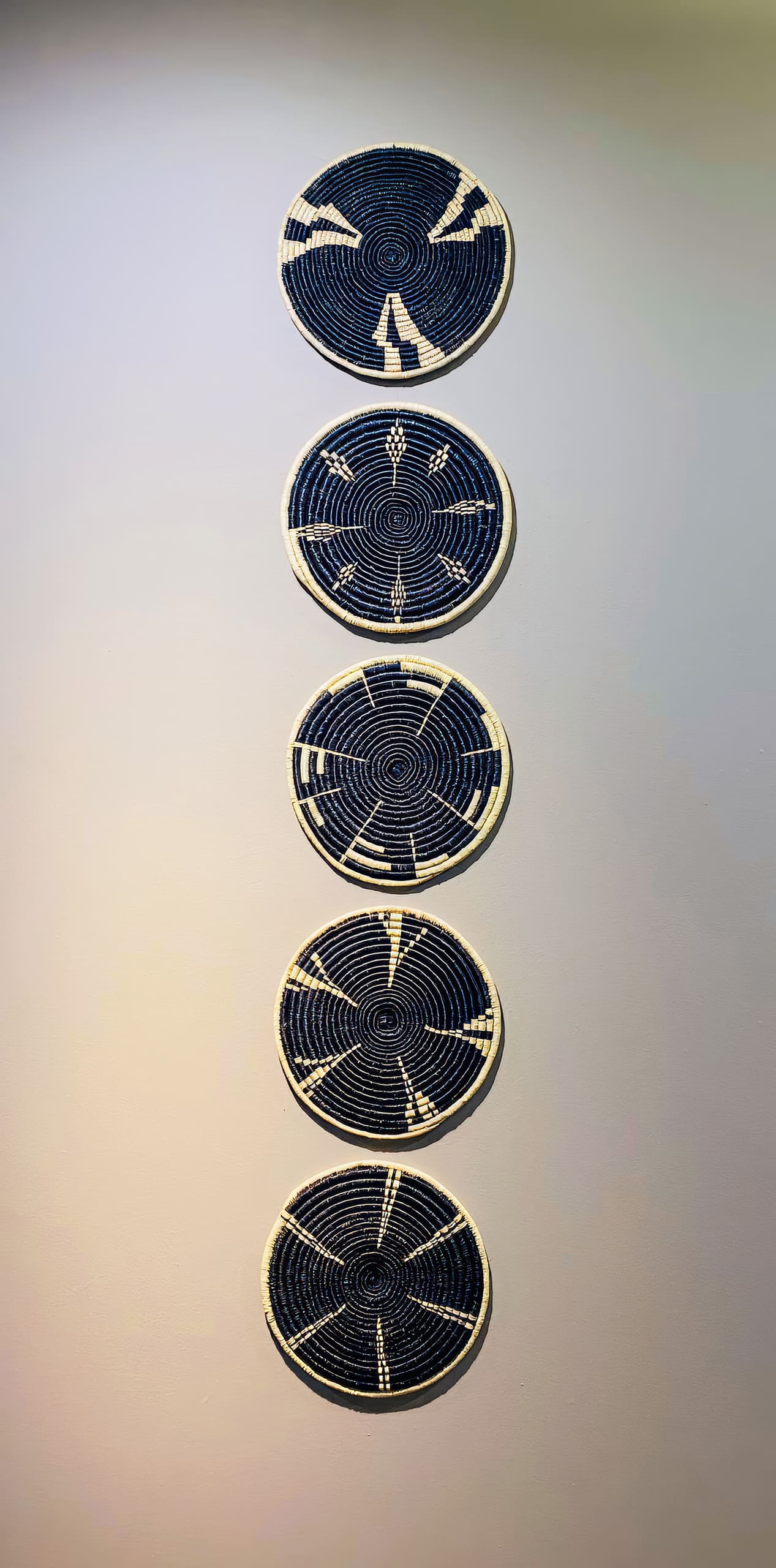 Six round, woven baskets with black and white geometric patterns are vertically aligned on a plain, light-coloured wall. Each basket features a unique design with concentric circles and pointed shapes, creating a visually striking column display.