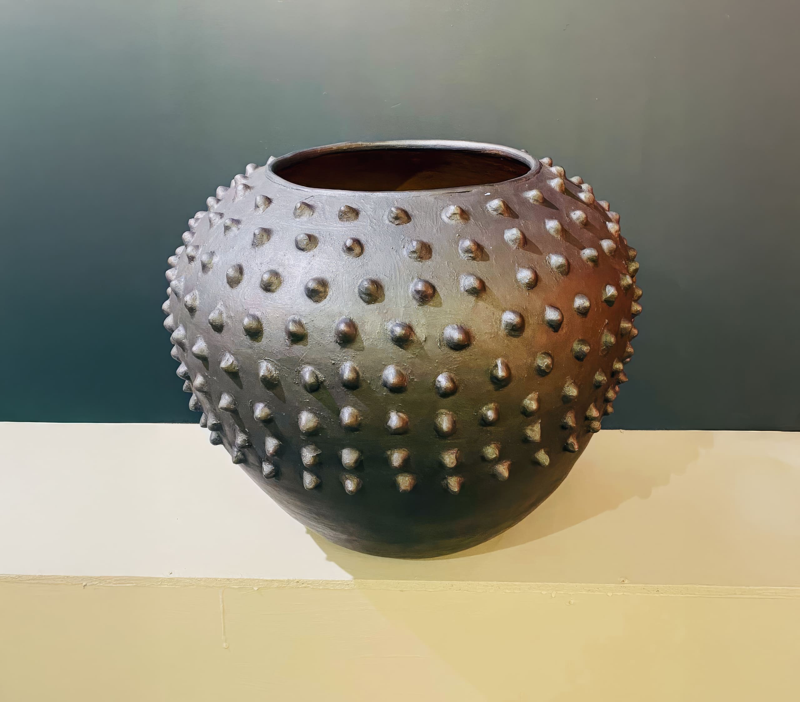 A round, dark ceramic vase sits on a light surface against a muted background. The vase features a unique design with evenly spaced, raised cone-shaped studs covering its exterior, giving it a textured and distinctive appearance.