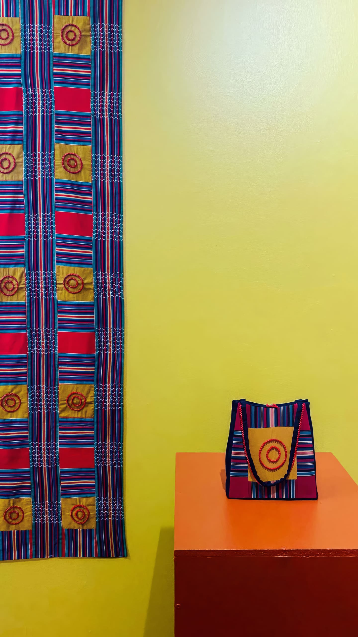 A brightly patterned fabric with red, yellow, and blue stripes and circular designs hangs on a yellow wall. Next to it, a matching handbag sits on an orange plinth, displaying a similar circular motif and colour scheme.