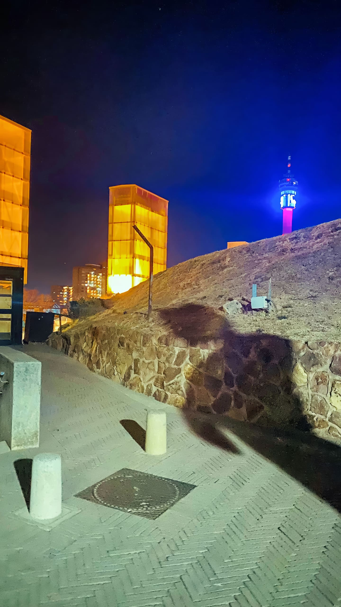 A nighttime scene with golden-lit glass towers on the left, a stone retaining wall, and a hill. A bright blue-lit tower rises at the right. The ground is paved, with bollards, benches, and a large shadow cast across the path. City buildings glow in the background.