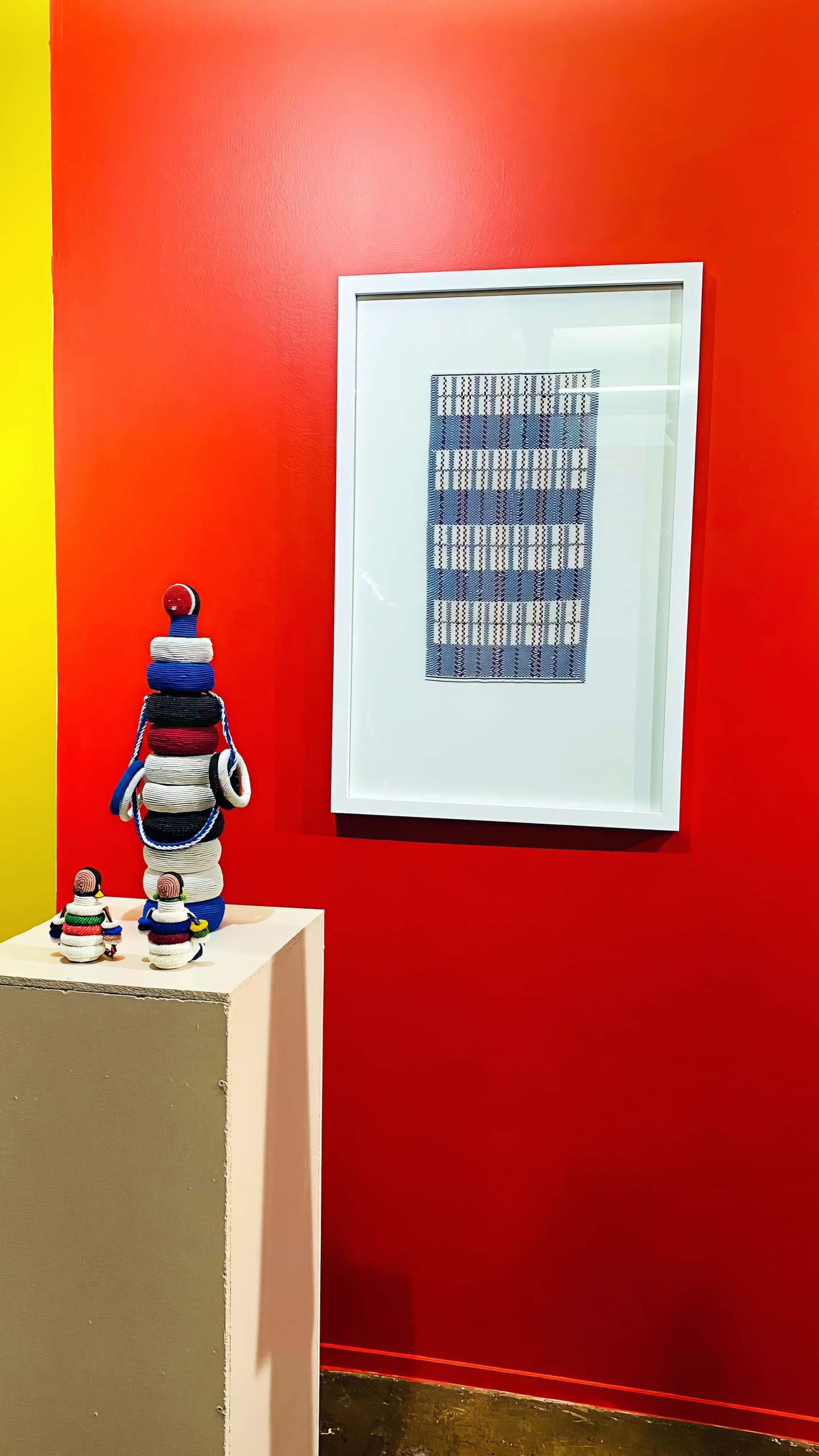 A framed textile art piece with a blue and white checked pattern hangs on a bright red wall. In front, a plinth displays three stacked, colourful, round soft sculptures, with two smaller similar sculptures beside it. The left wall is yellow.