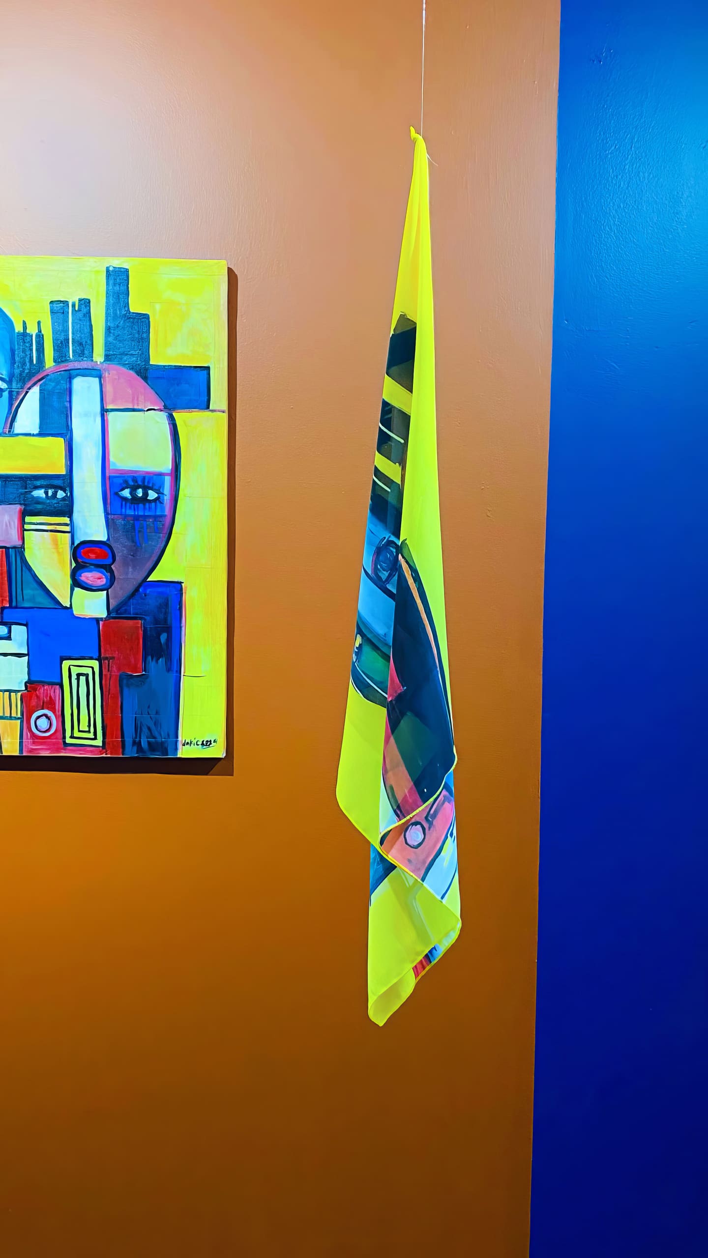 A colourful abstract painting featuring a geometric face hangs on an orange wall beside a bright yellow cloth with a similar pattern, draped from a hook. The wall has orange and dark blue sections divided vertically.