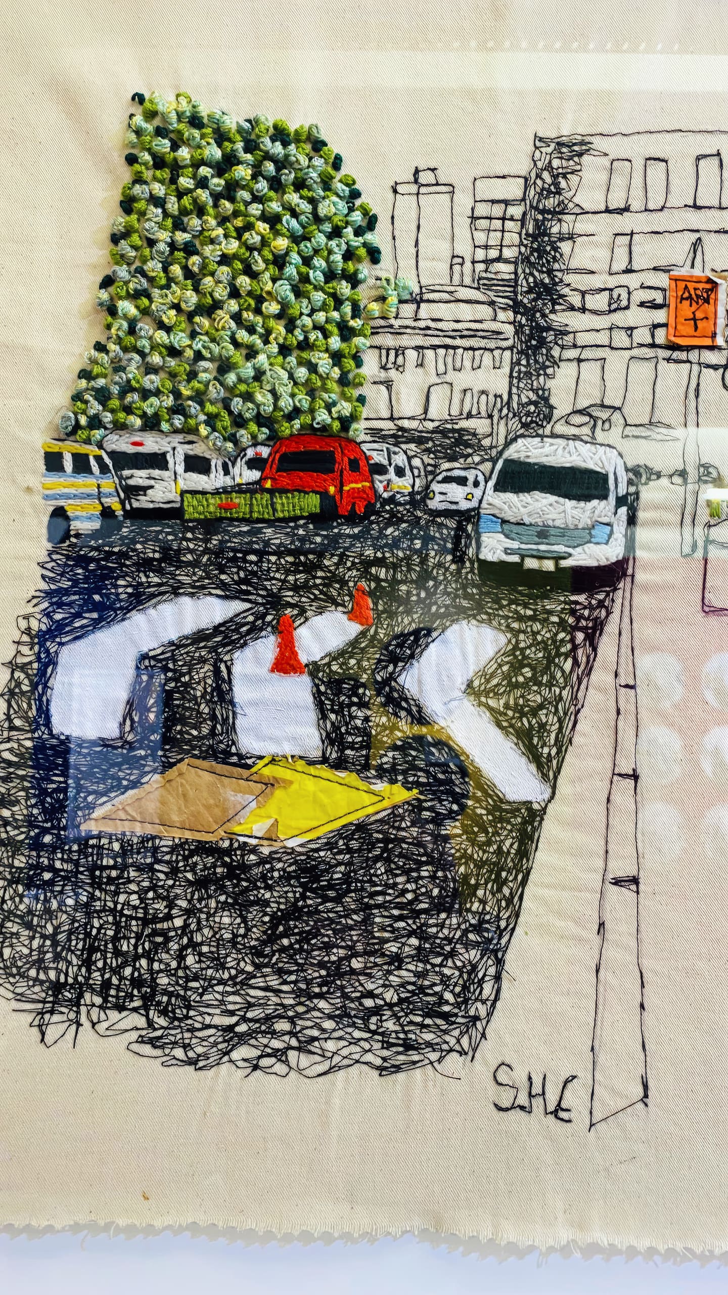 A textile artwork depicts a city street with stitched outlines of buildings, cars, and road markings. The left shows a textured green tree made of knotted fabric, whilst the rest features detailed black thread drawings, including orange cones, arrows, and a few coloured vehicles.