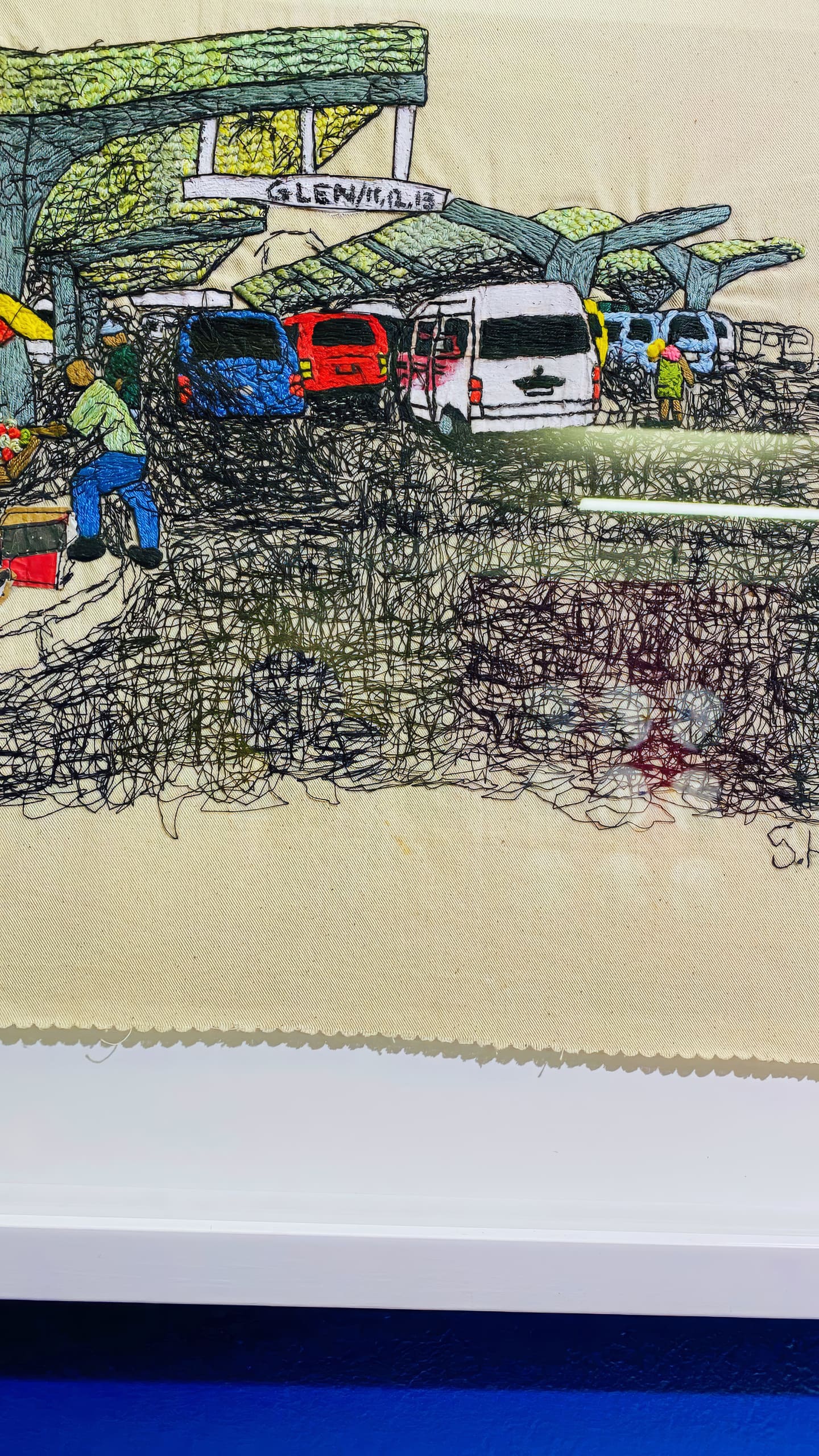 Textile artwork depicting a busy street scene with parked cars, people walking, and green awnings overhead. The sign above reads 