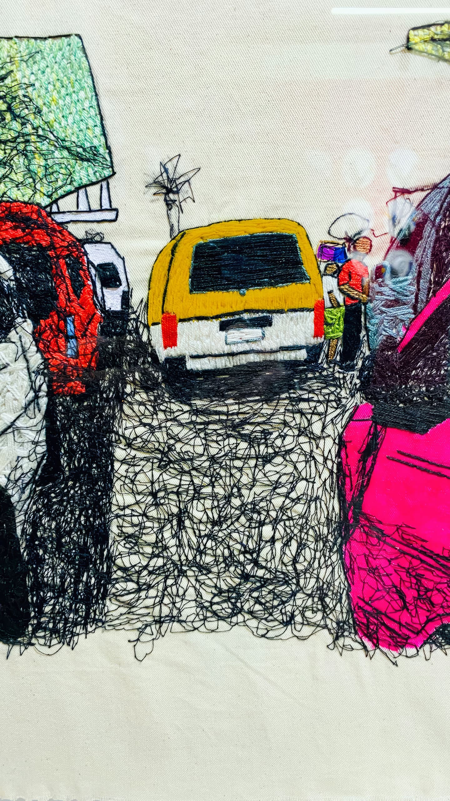Colourful thread embroidery depicts a street scene with cars, including a central yellow van, and two people in safety vests beside it. The textures are made with loose, overlapping stitches, adding depth and vibrancy to the urban setting.