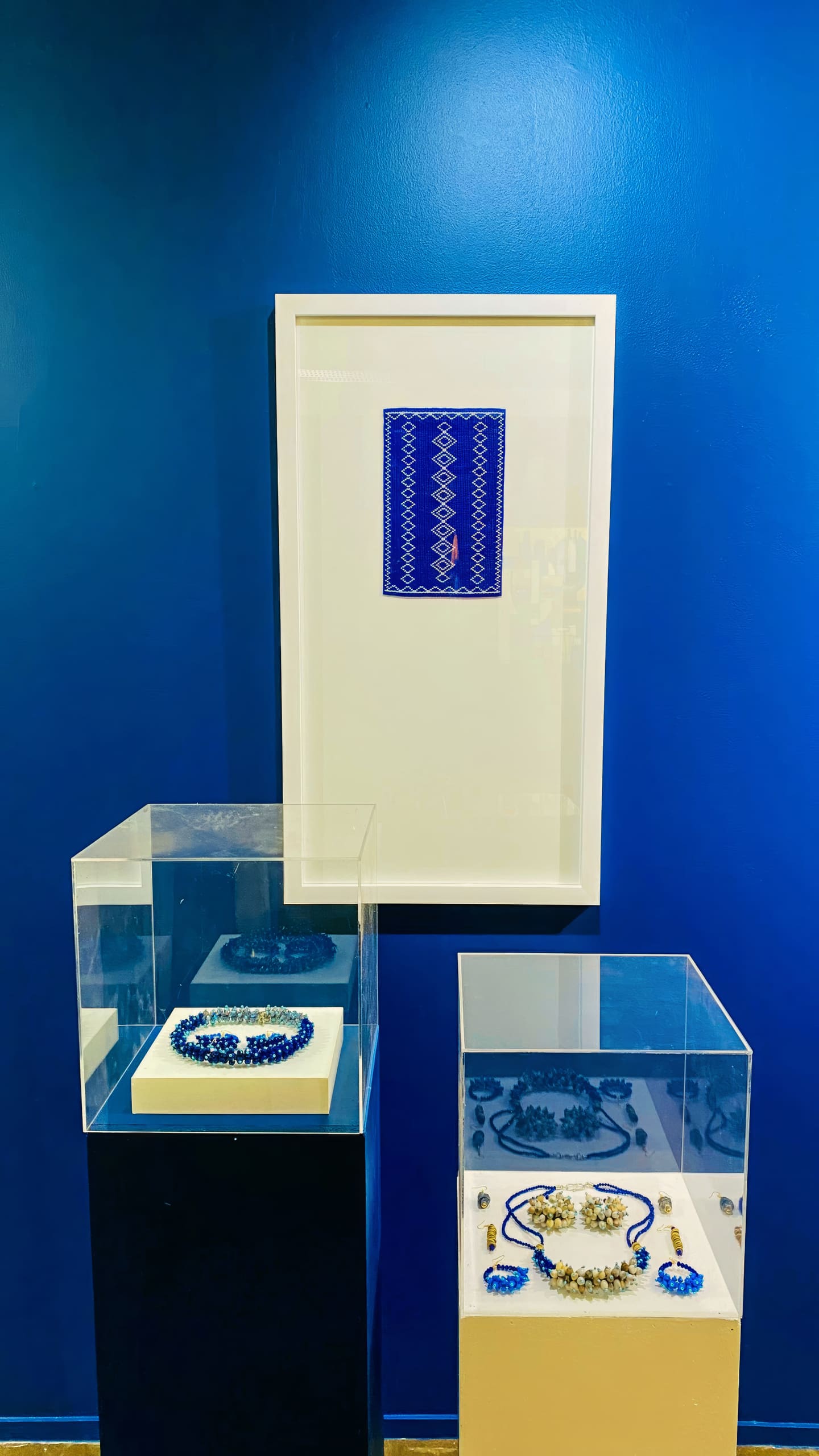 Jewellery display against a vivid blue wall: two glass cases on pedestals hold ornate necklaces and earrings, predominantly in blue and gold tones. Above them, a framed textile piece with a geometric blue and white pattern hangs on the wall.