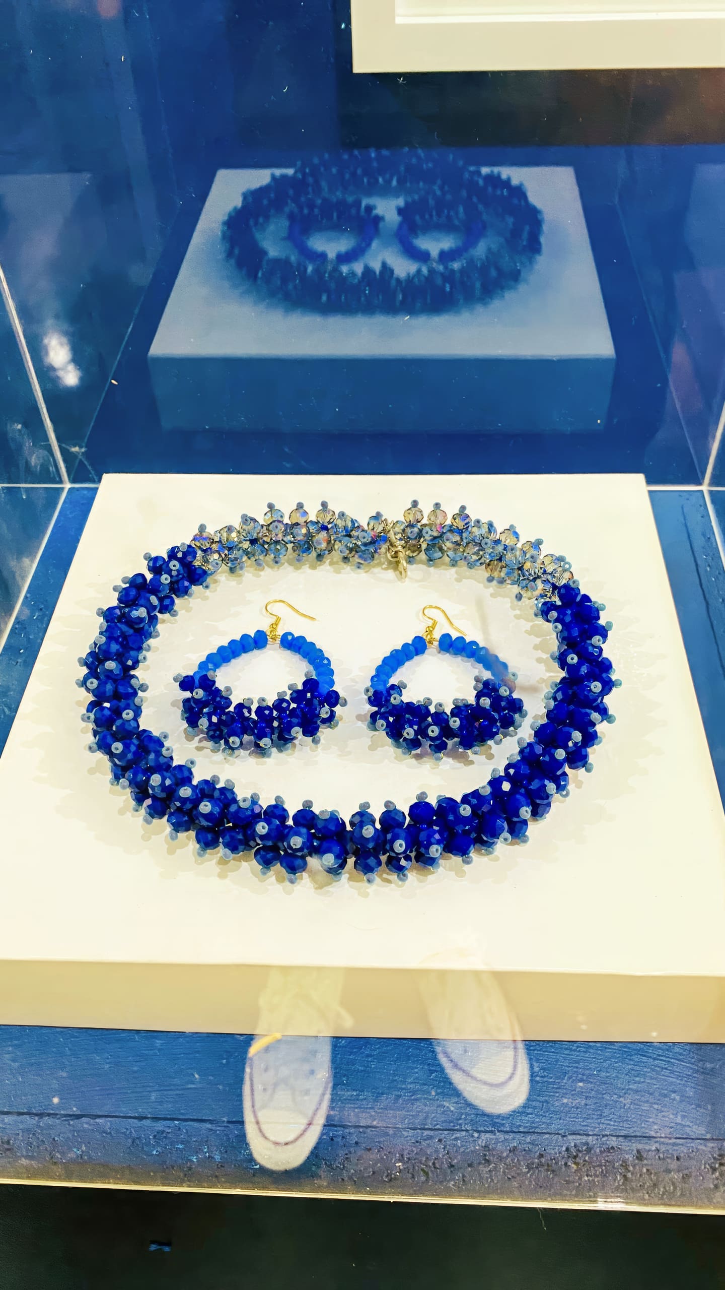 A set of blue beaded jewellery, including a necklace and matching hoop earrings, is displayed on a white platform inside a glass case. The beads are deep blue with some lighter blue accents. The jewellery’s reflection is visible in the case behind it.