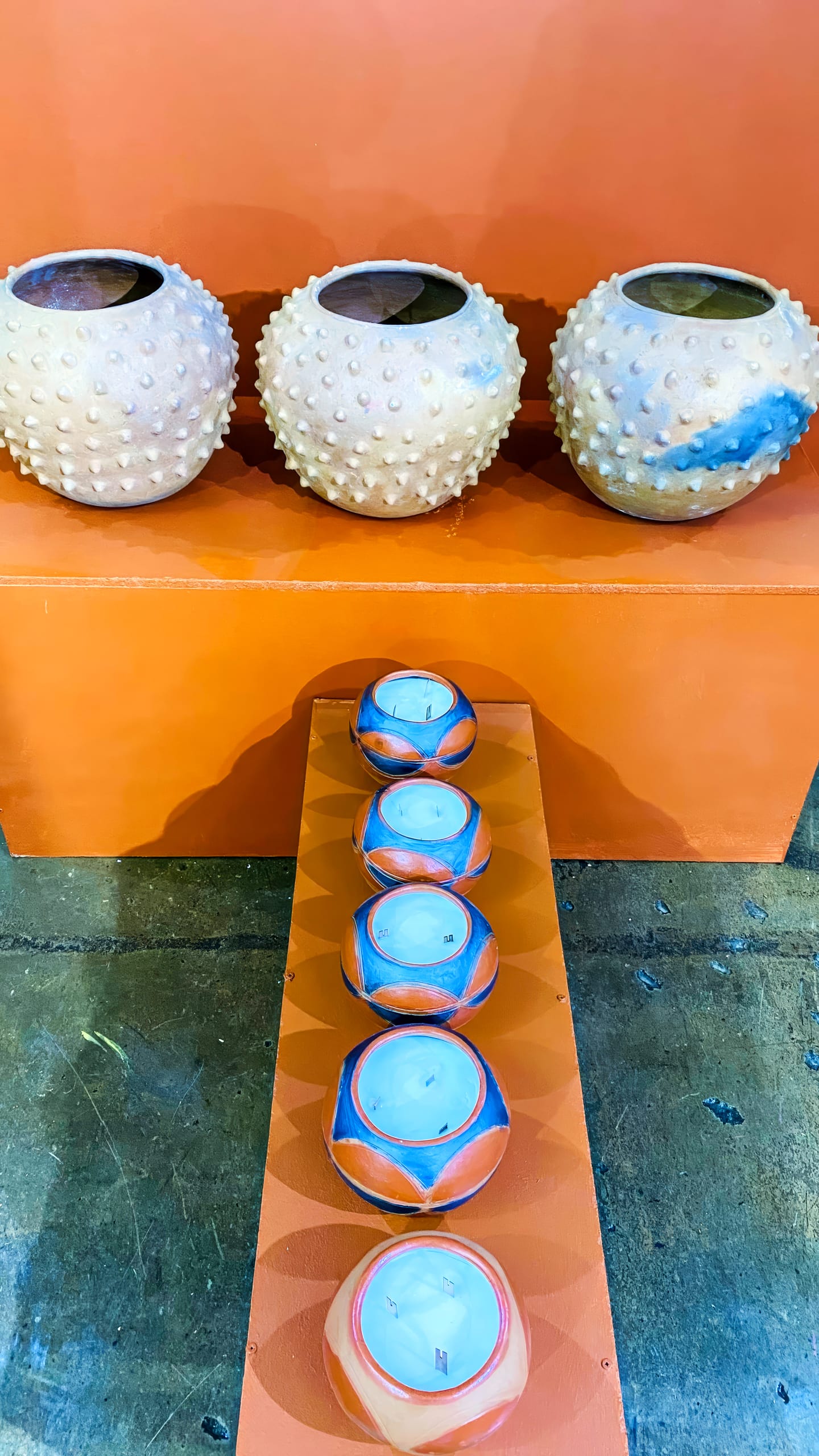 Three round, beige, spiked ceramic vases from the BASA Asiwele collection sit on an orange shelf. Below, a row of six orange-and-blue candle holders with white candles are arranged neatly against a concrete floor background.
