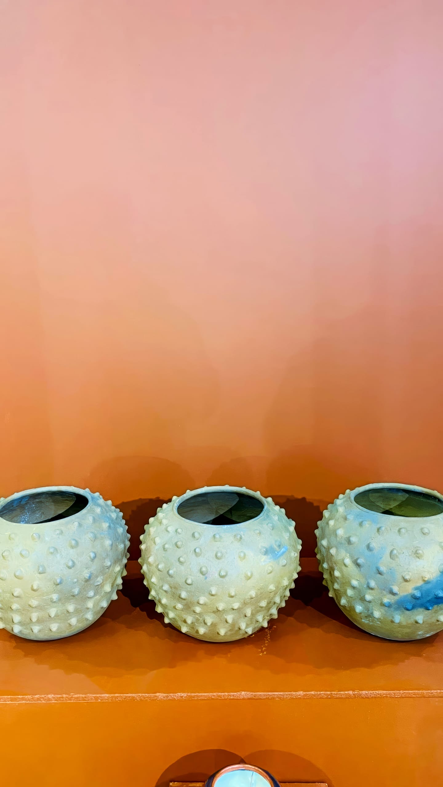Three round, beige ceramic pots with raised, bumpy textures are displayed side-by-side on an orange shelf against a peach-coloured wall. Inspired by BASA Asiwele: Crossing the Divide, the minimalist arrangement evokes unity and balance.