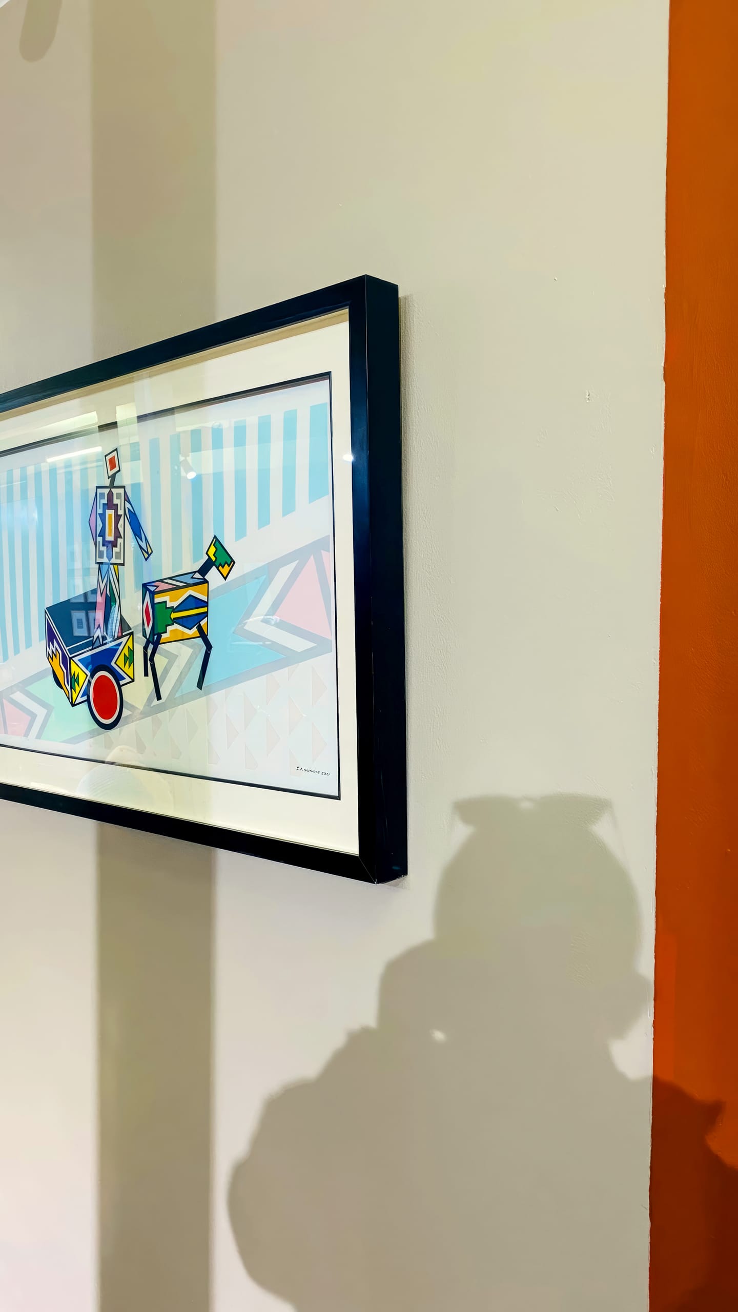 A framed abstract artwork, inspired by BASA Asiwele, hangs on a light-coloured wall next to an orange vertical stripe. The painting features geometric shapes and figures in blue, yellow, and pink tones. A shadow of a person holding a mobile appears in the foreground.