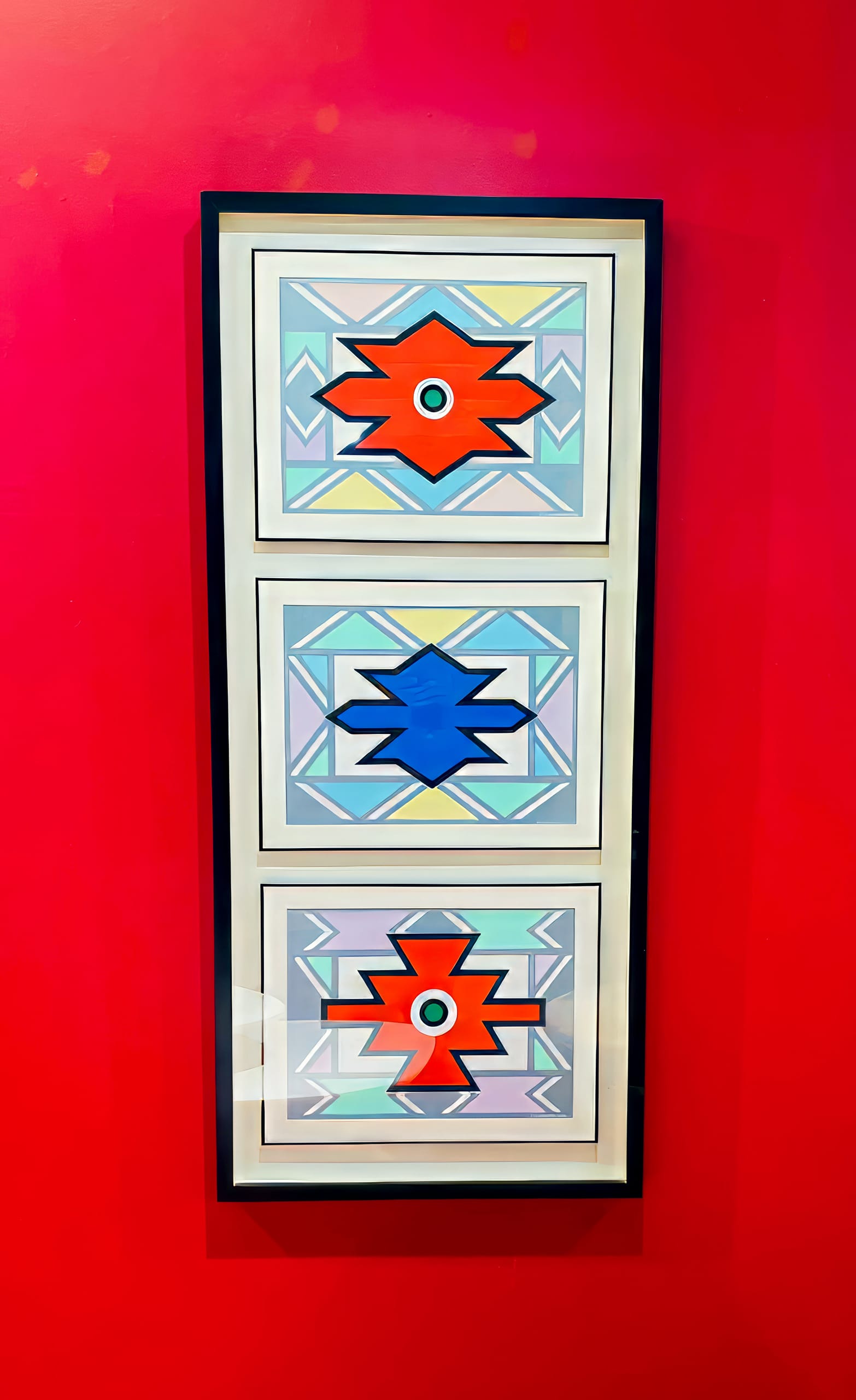 A framed artwork, titled Crossing the Divide, features three geometric starbursts—red, blue, and red—set against pastel backgrounds with turquoise and grey tones. It hangs on a bright red wall, making a vivid BASA Asiwele statement.