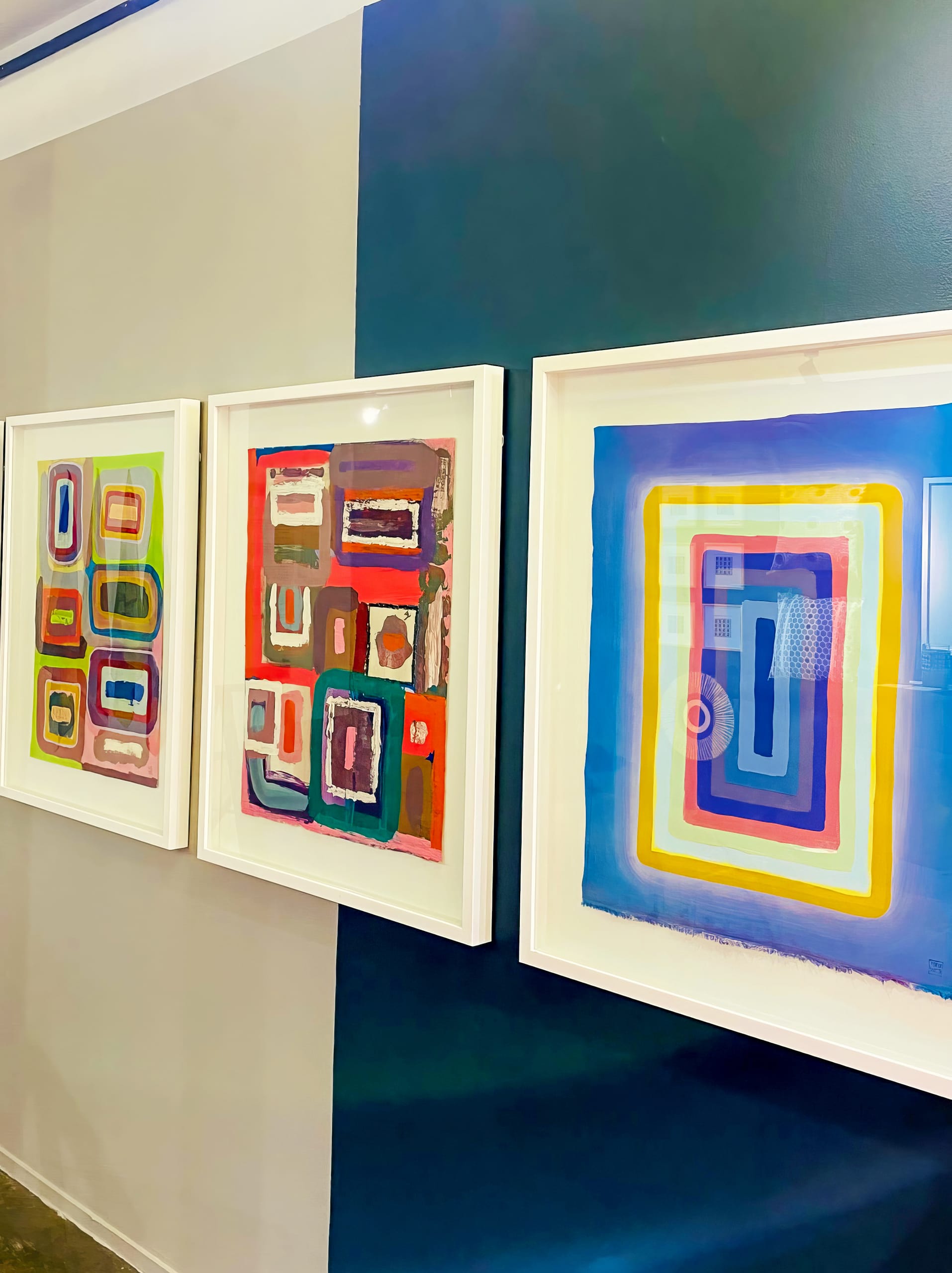 Three framed abstract paintings, inspired by BASA Asiwele, with vibrant overlapping rectangles hang side by side on a wall painted in vertical blocks of grey and dark teal, featuring striking blues, reds, yellows, greens, and purples.