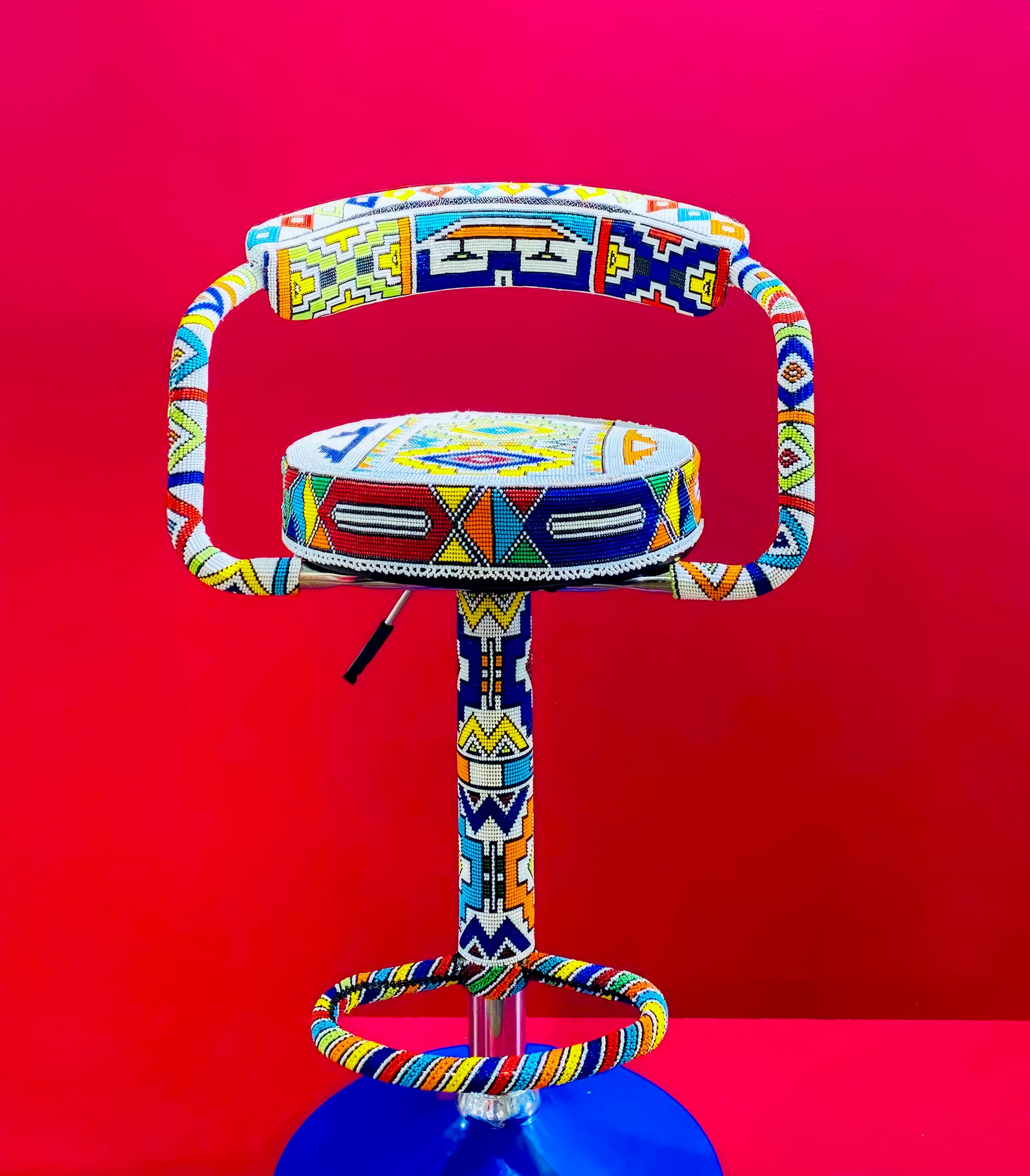 A colourful barstool decorated with intricate beadwork in geometric patterns, inspired by BASA Asiwele, stands against a vibrant pink background. The seat, backrest, and legs are all adorned with detailed designs in yellow, blue, red, green, and white beads.