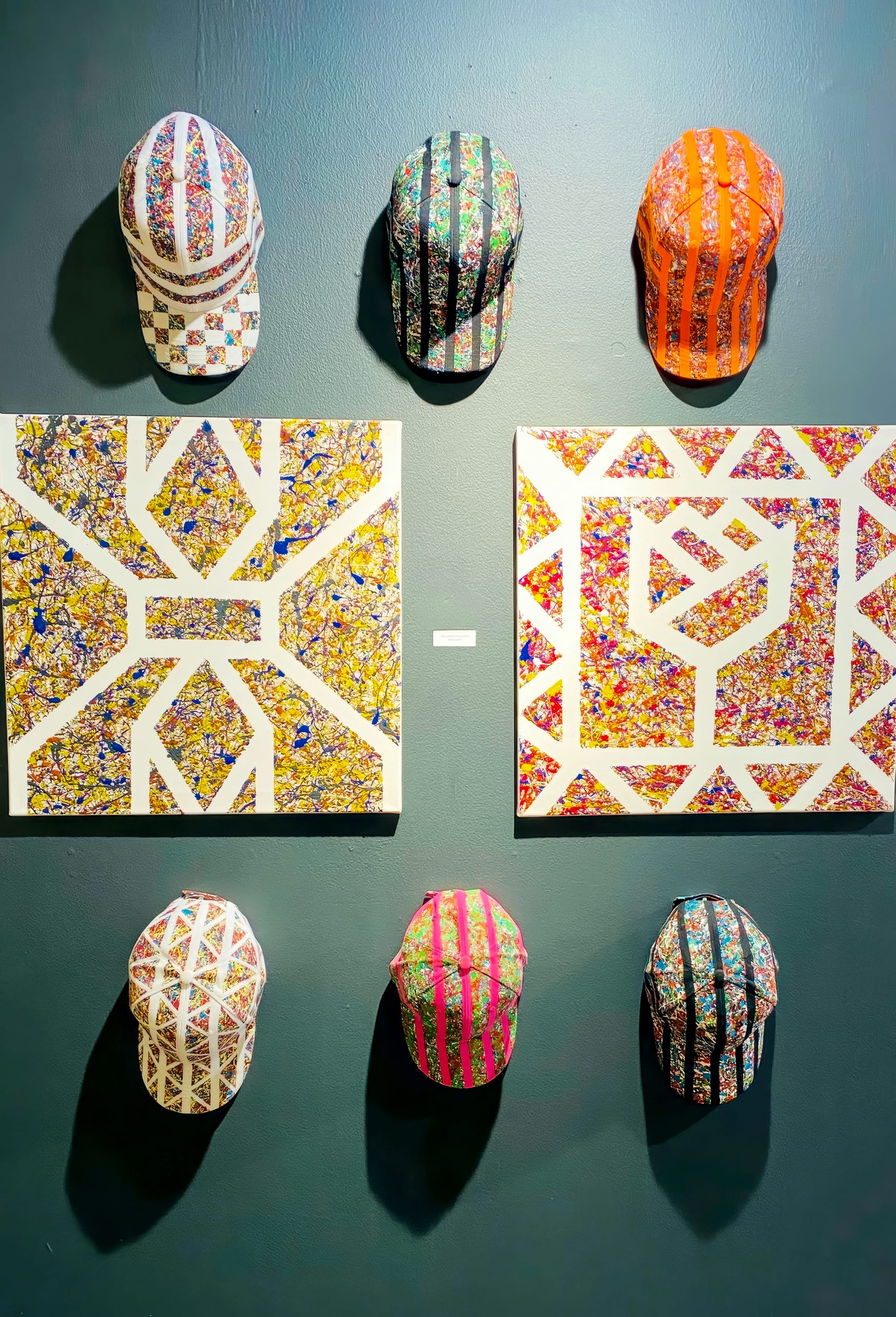Six colourful, patterned caps are displayed on a dark wall around two abstract, geometric artworks. Titled 