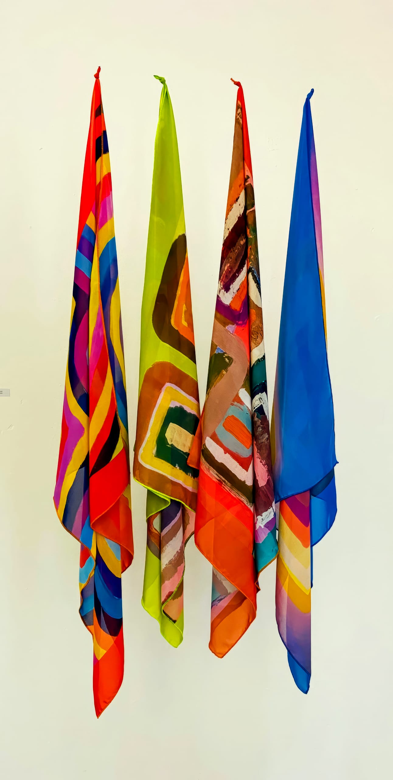 Four vibrant, patterned fabric scarves hang vertically against a white wall. Part of the BASA Asiwele collection, each scarf features bold, abstract designs and vivid colours—Crossing the Divide between art and fashion in a striking display.
