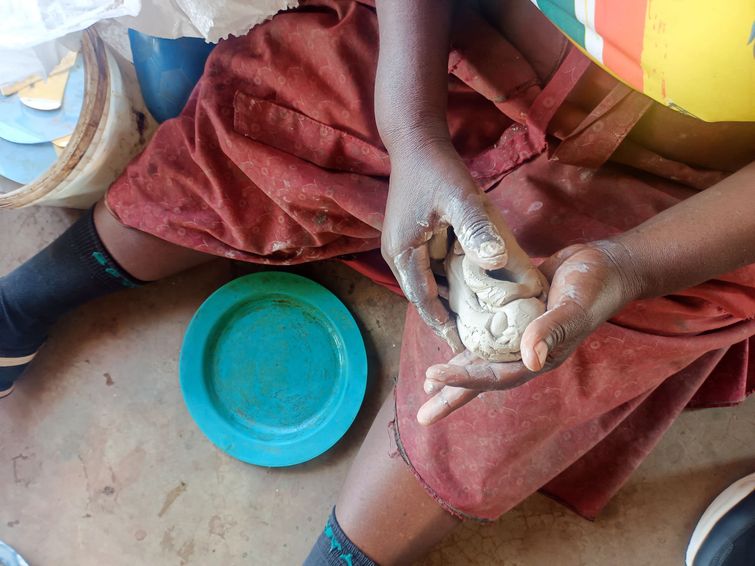 A person wearing a red skirt, striped shirt, and dark socks shapes a lump of clay with their hands whilst sitting on the floor. A blue plastic plate and a white basket are placed nearby on the ground.