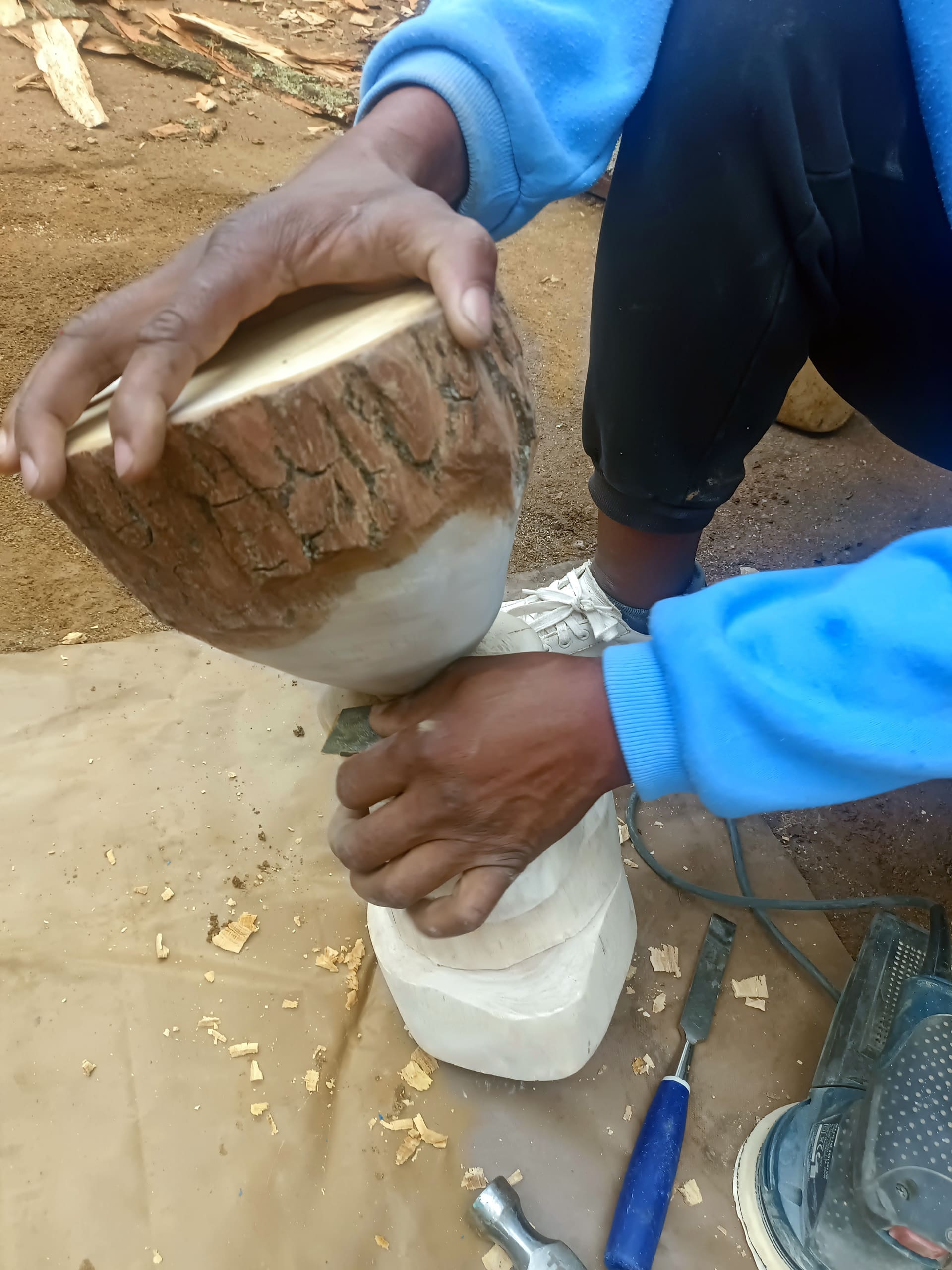 A person wearing a blue sleeve shapes a wooden object with both hands, holding it steady on a work surface scattered with wood shavings. Part of the wood retains bark, and carving tools are visible nearby. The person's white shoe and brown ground are also seen.